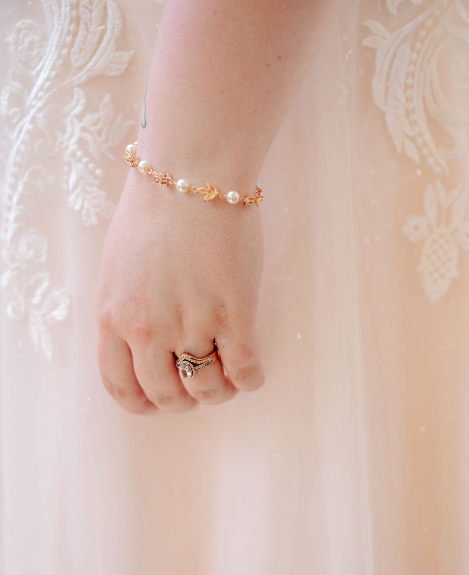 Bride wearing a delicate gold and pearl bracelet with stacked rings on her hand against a white lace wedding gown.