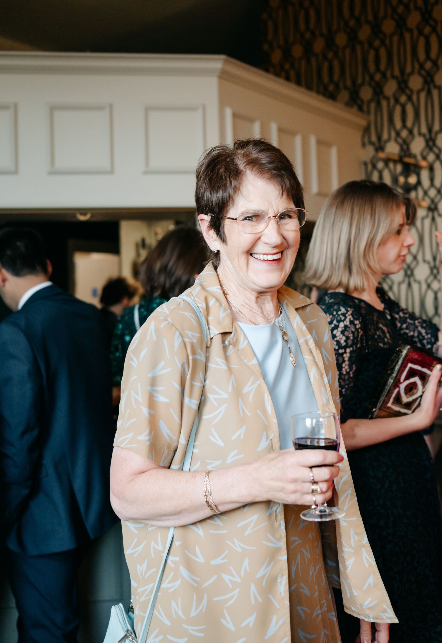 Guest in beige patterned outfit enjoying wine at an elegant indoor reception.