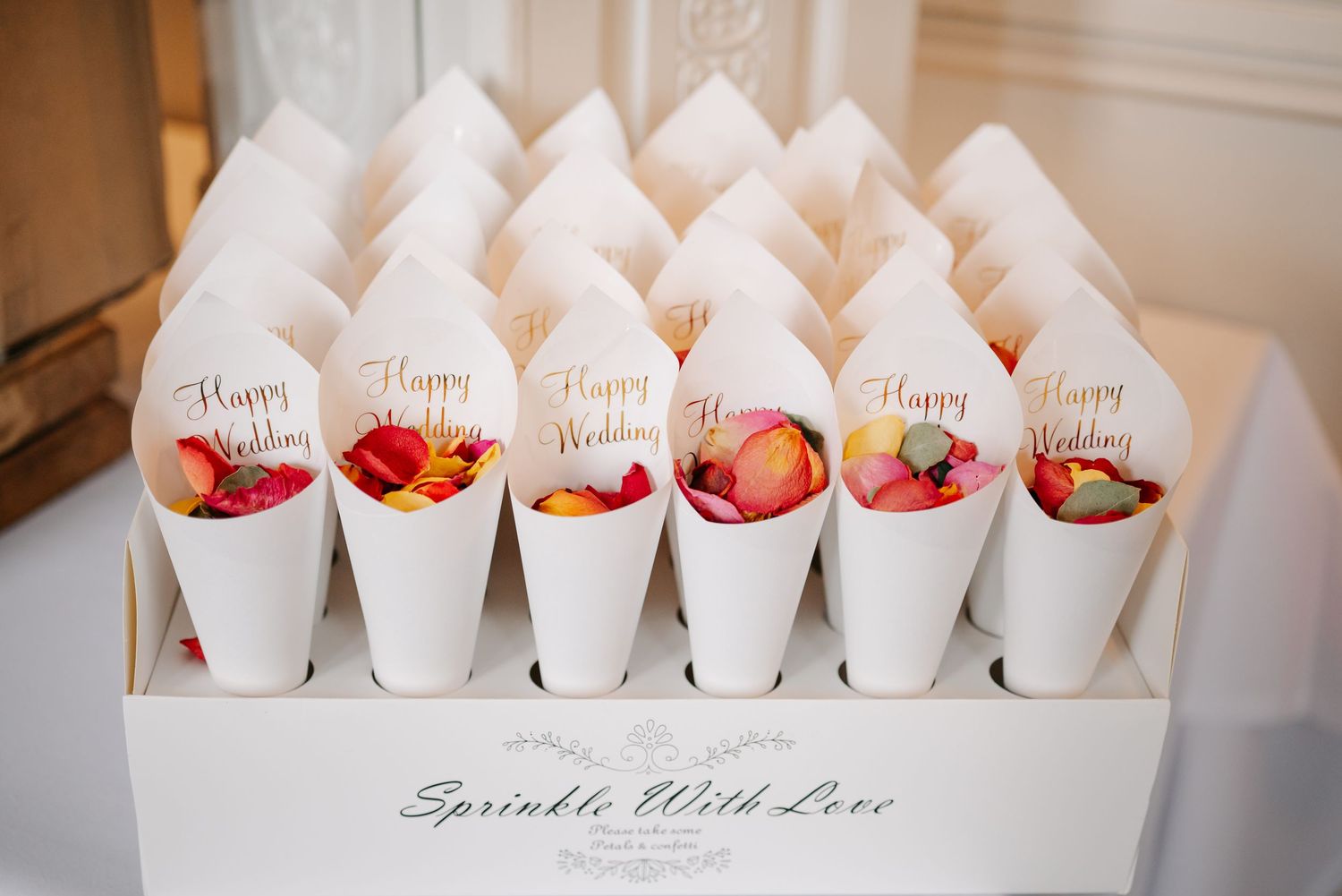 Rows of white paper cones filled with rose petals are displayed on a table for a wedding ceremony.