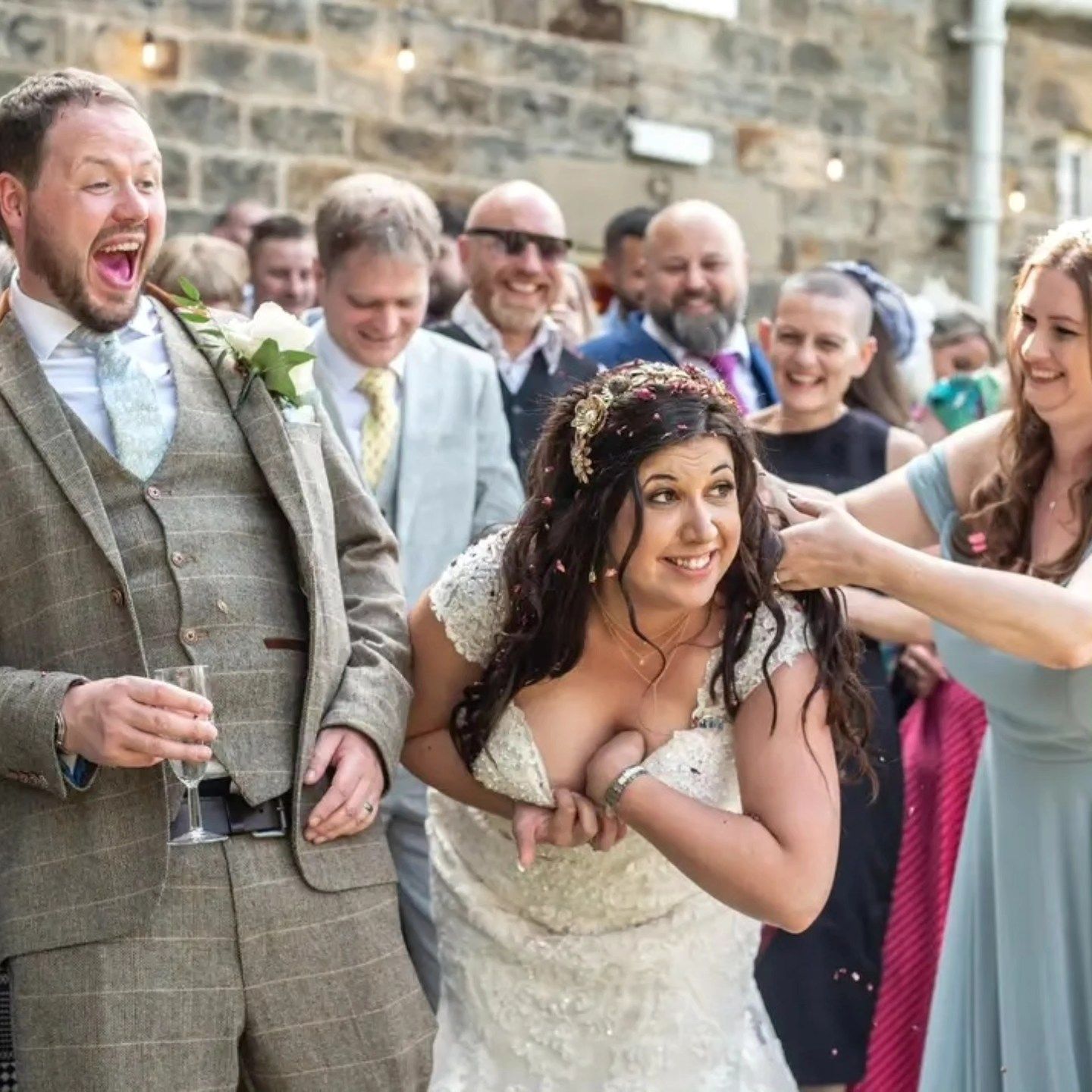 Wedding guests toss confetti over newlyweds during outdoor celebration with brick wall backdrop.