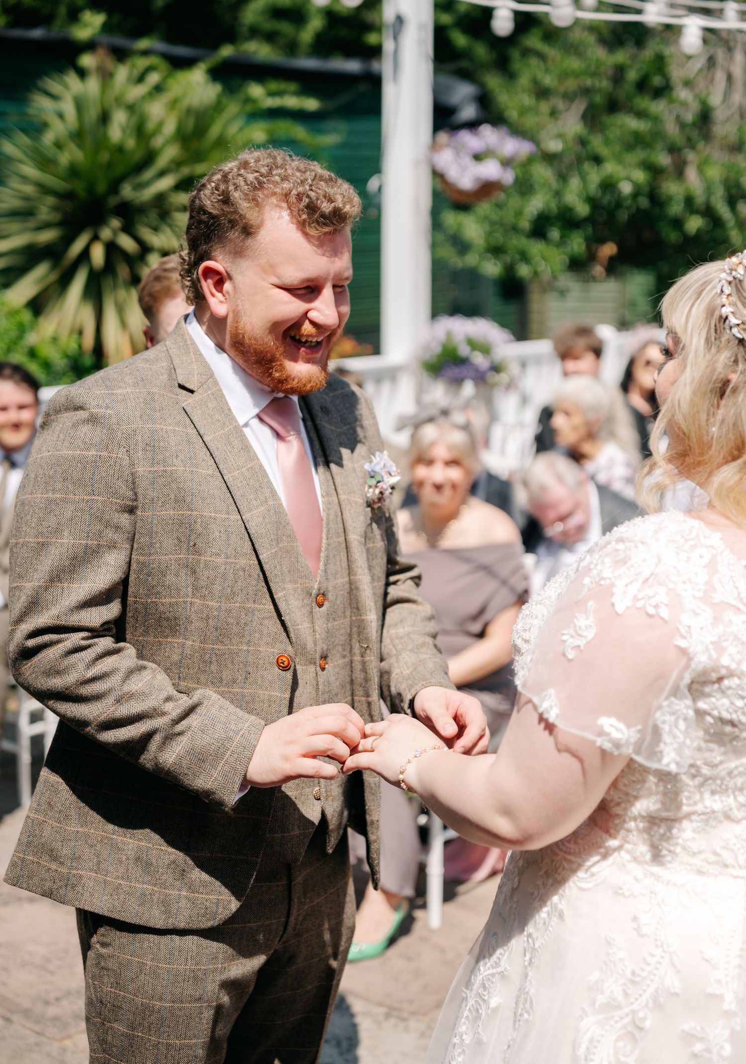 Groom smiling as bride places wedding ring on his finger during sunny outdoor ceremony.