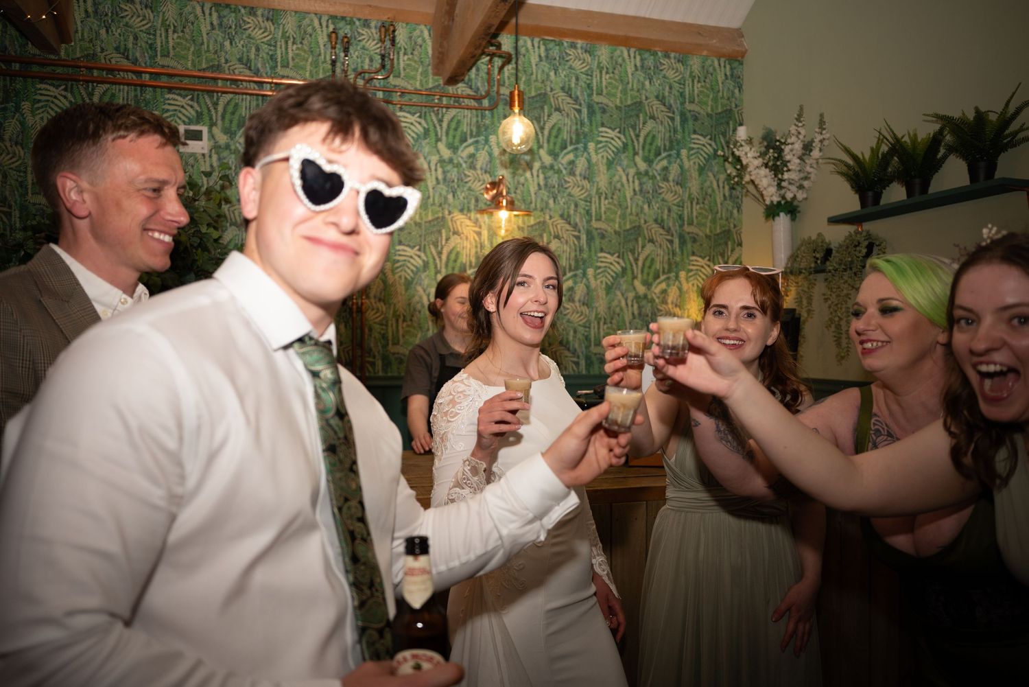 Wedding reception guests celebrate with drinks and sunglasses in a festive indoor venue.