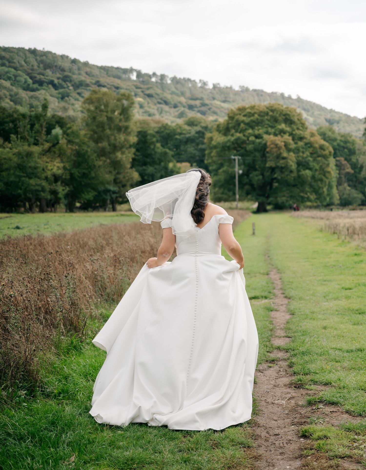 A flowing white wedding dress billows in the breeze as someone walks along a countryside path near green hills.