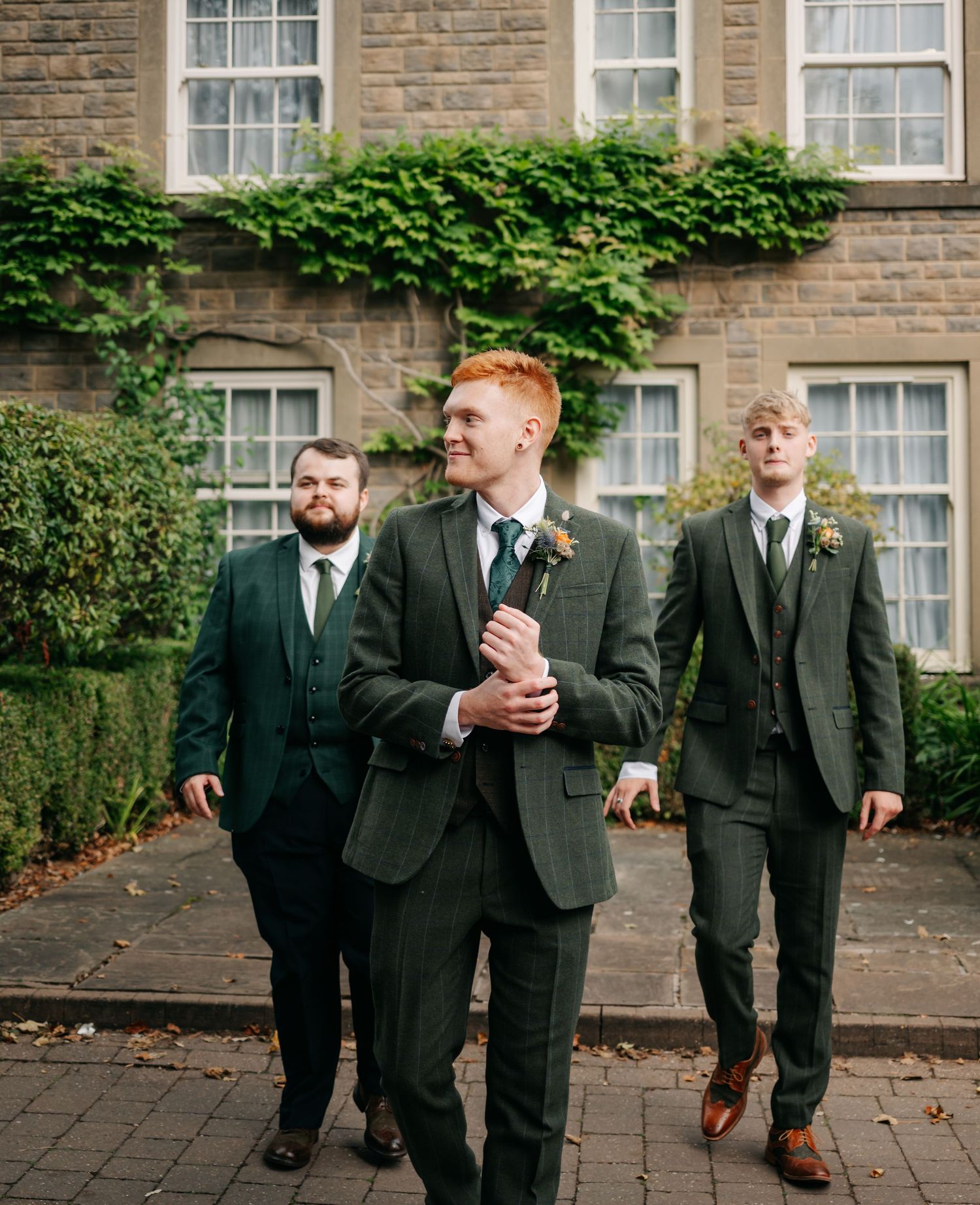 Wedding party members in dark suits walking outside a stone building with ivy-covered walls.