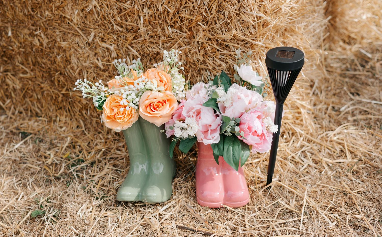 Colorful rain boots filled with fresh flowers arranged as rustic wedding decor on hay-covered ground.