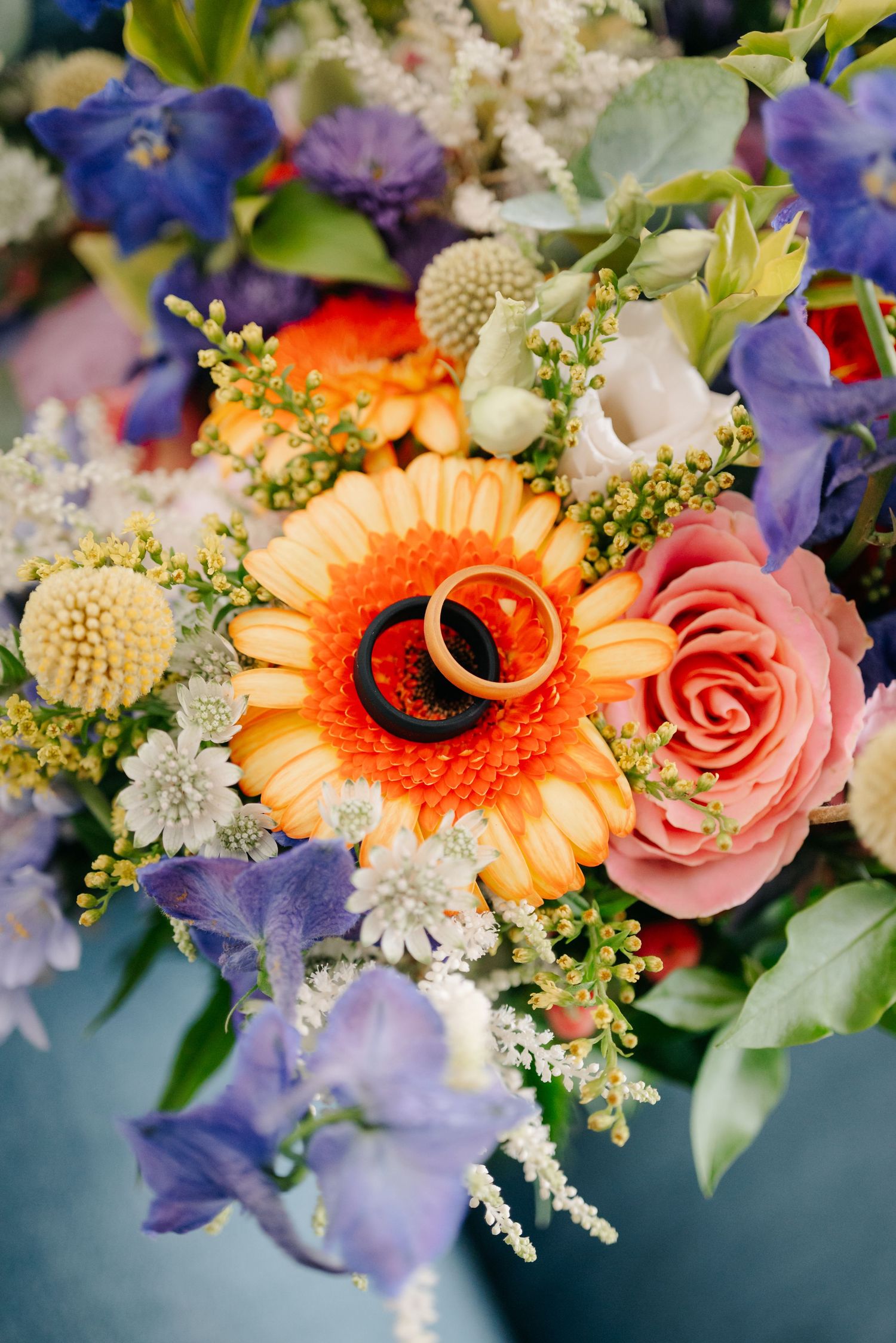 Vibrant floral arrangement featuring orange gerbera daisies, pink roses, purple irises, and white accents against a blue background.