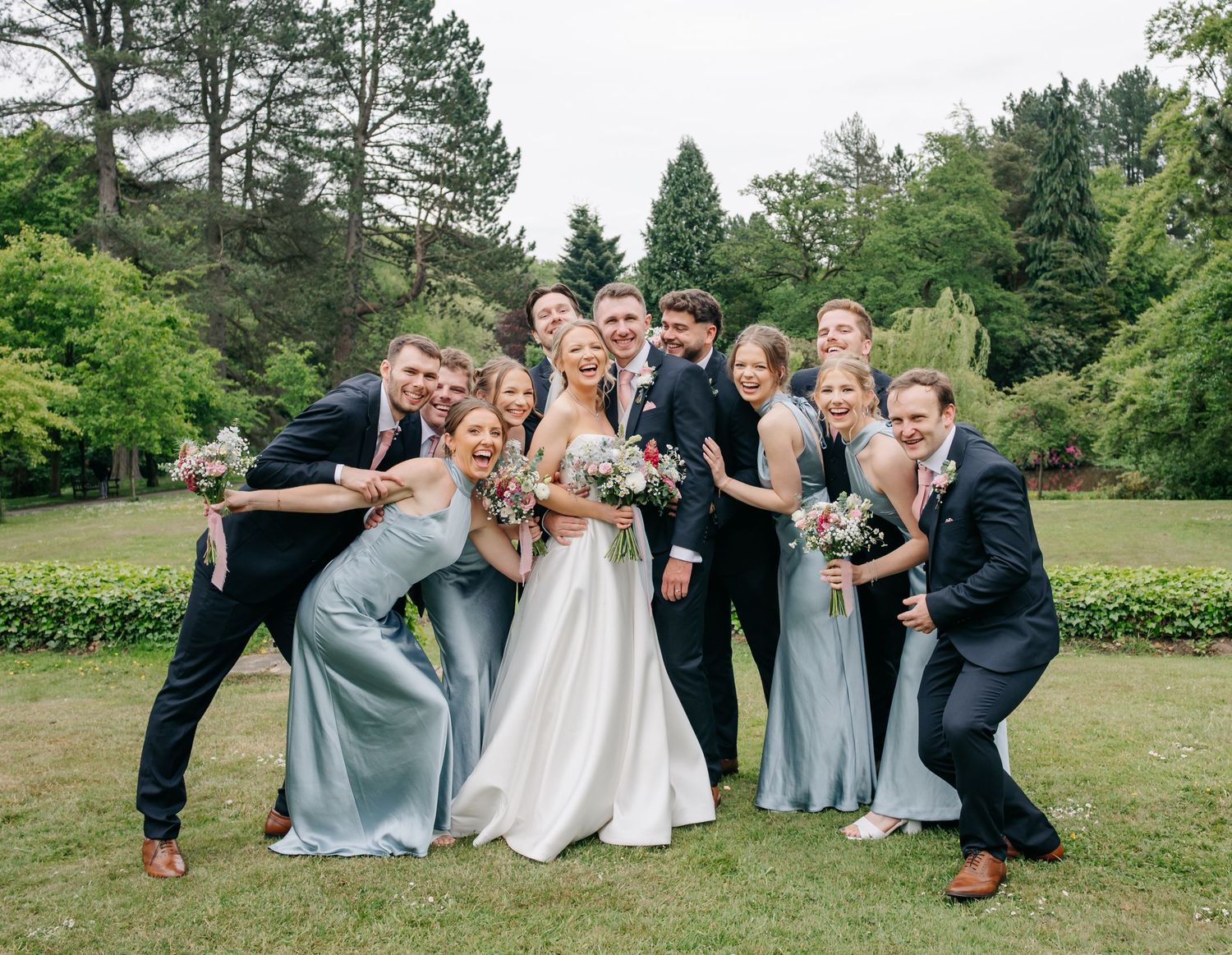 Wedding party poses playfully in a garden setting wearing blue dresses and dark suits for fun group photos.