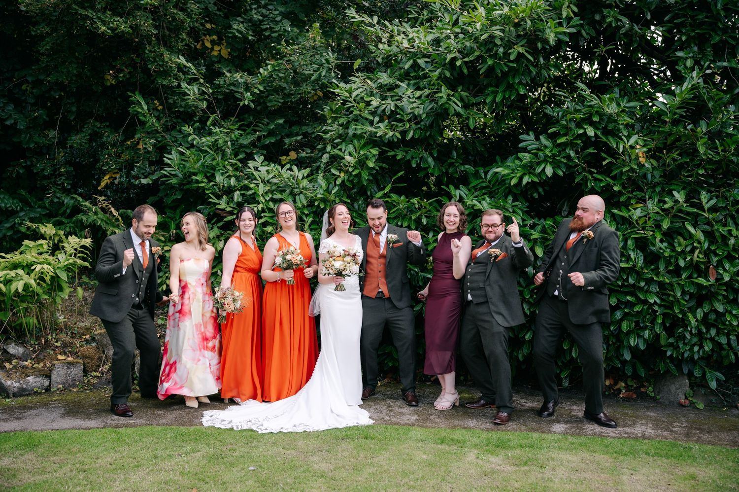Bride in white gown with bridesmaids in coral and pink dresses and groomsmen in dark suits cheering together on a lawn before green hedges.