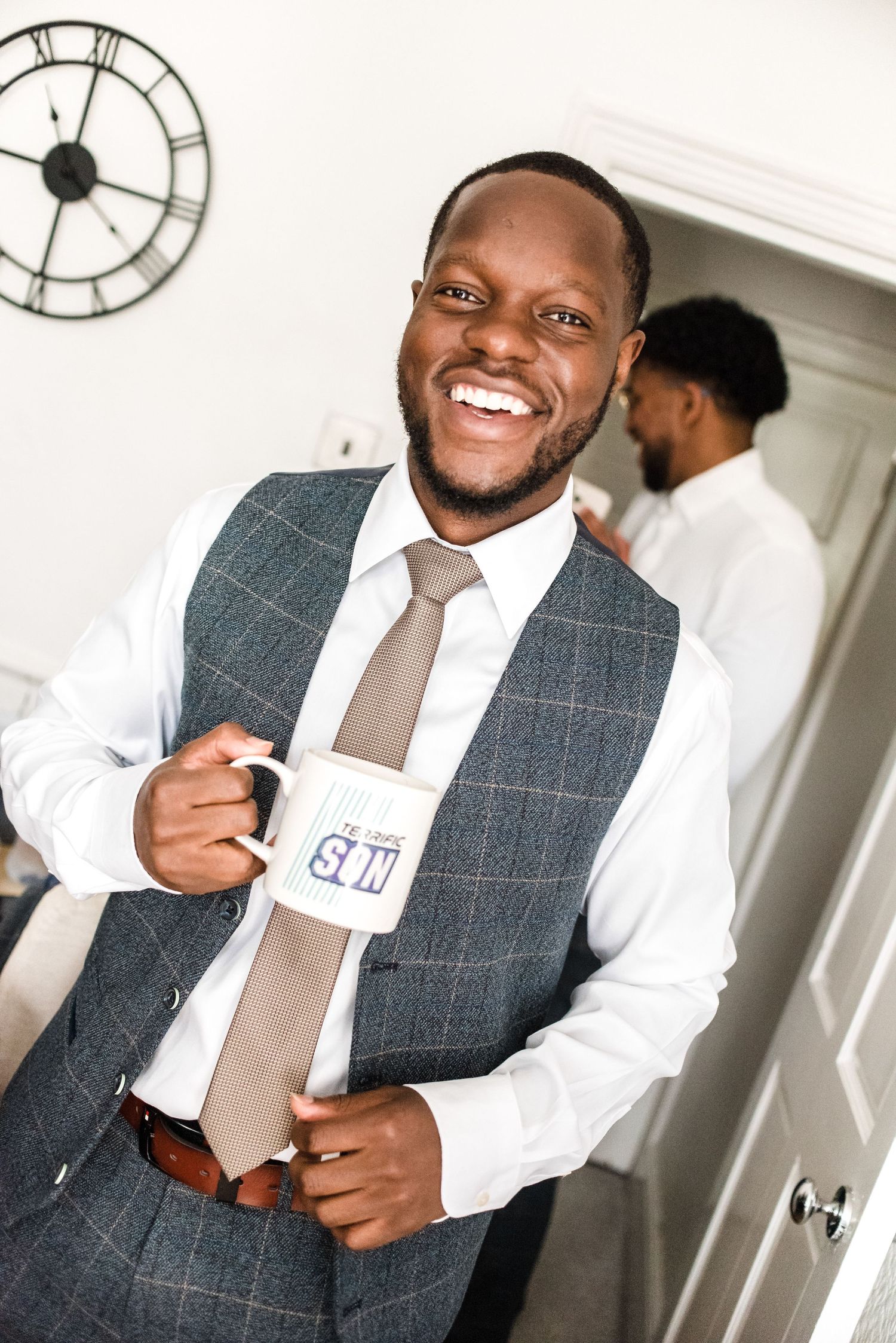 A guest in formal attire holds a coffee mug while smiling warmly in an indoor setting with a clock on the wall.