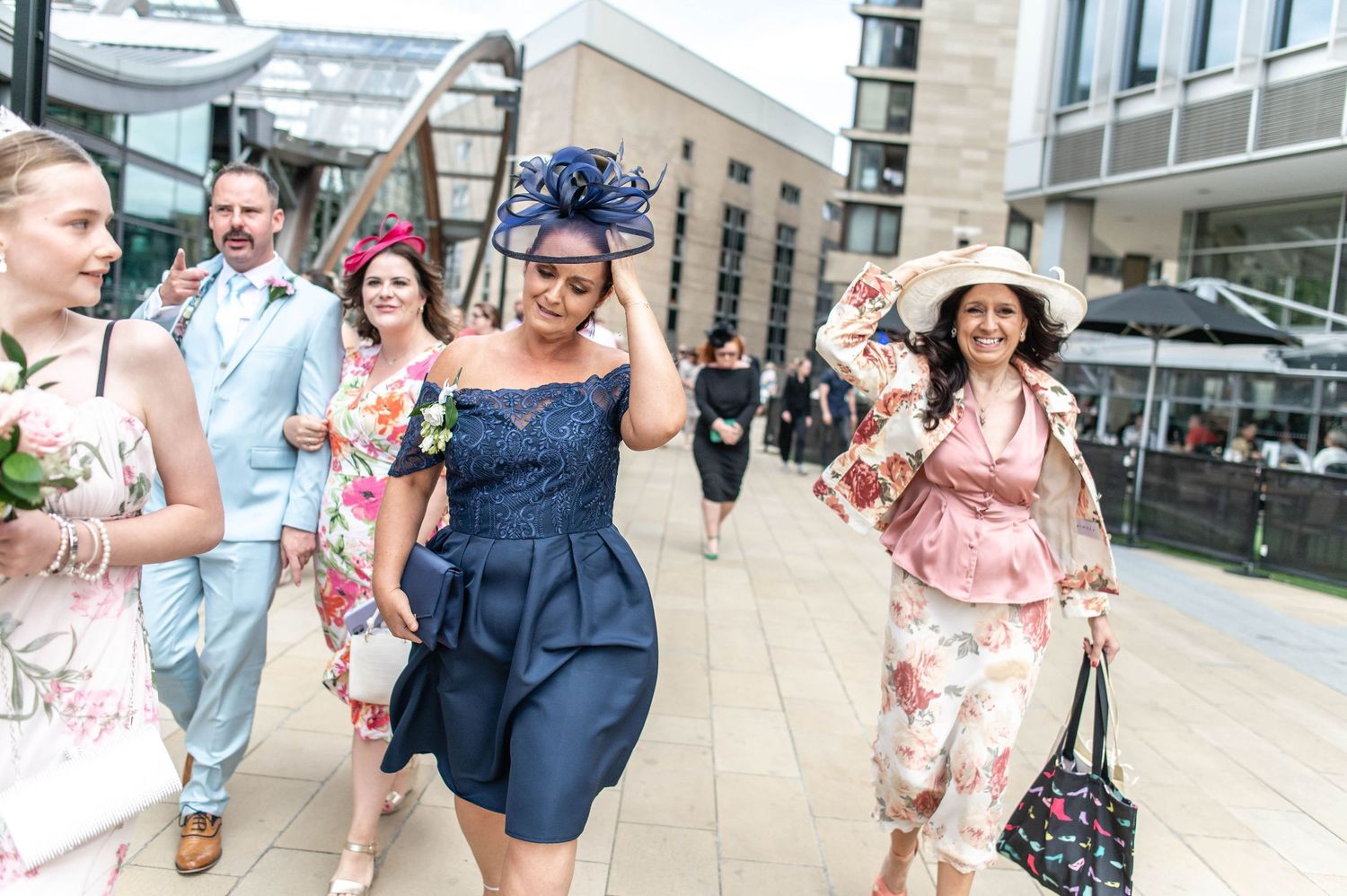 Wedding guests in formal attire and fascinators walking and laughing together on a city street.