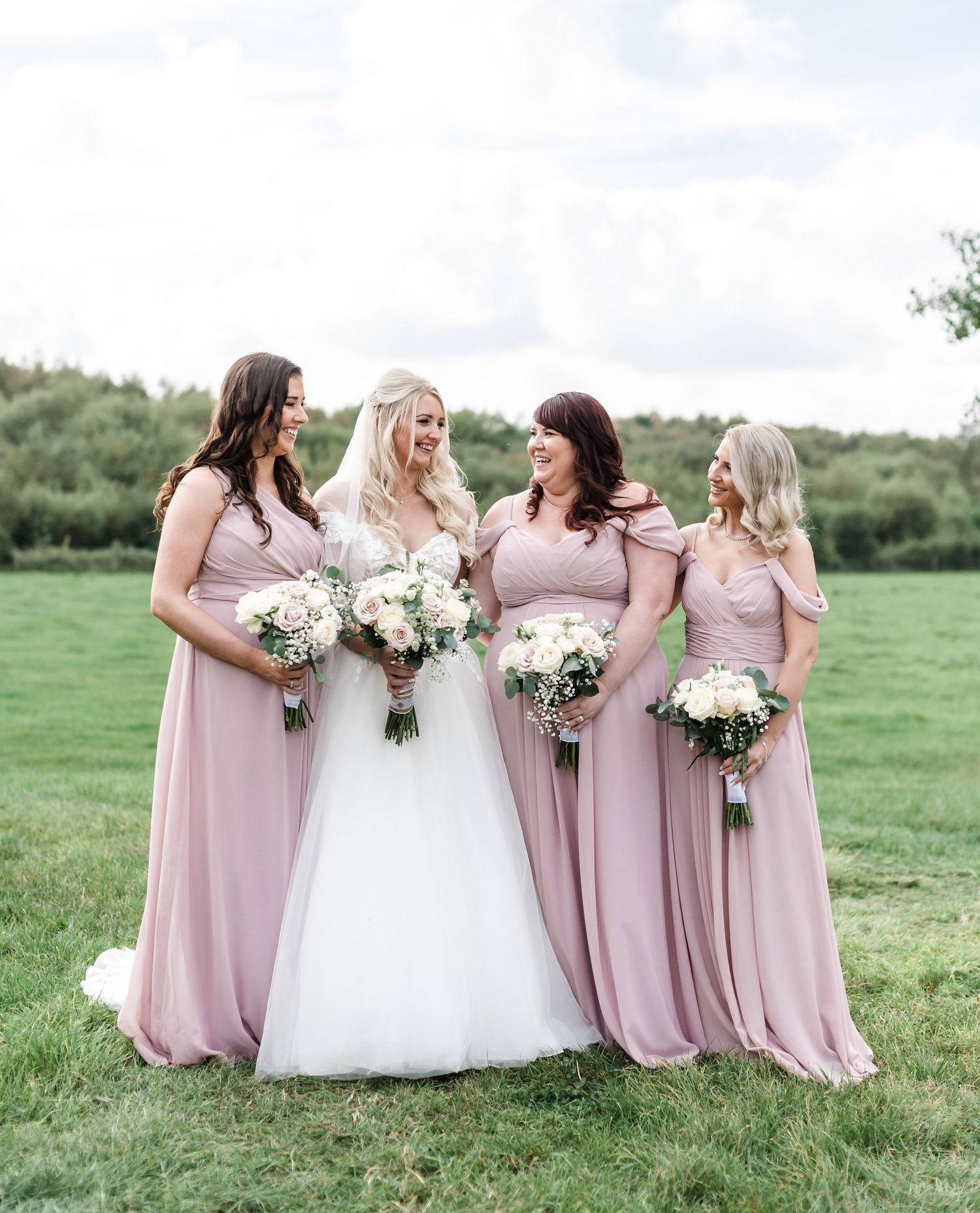 Group in pink bridesmaid dresses and white wedding gown pose together in a green field.