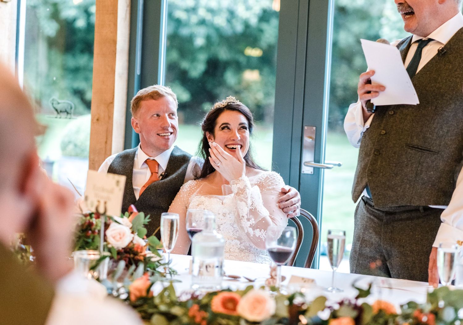 Indoor wedding reception with guests reacting to speeches at decorated table.