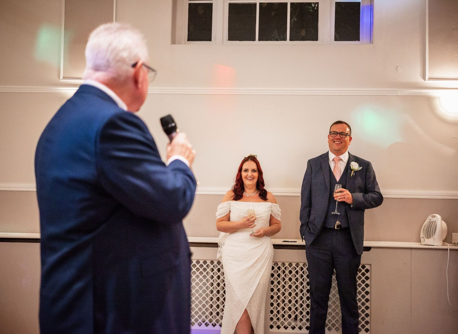 Wedding reception scene with couple listening to speech in white-walled venue with decorative lighting.
