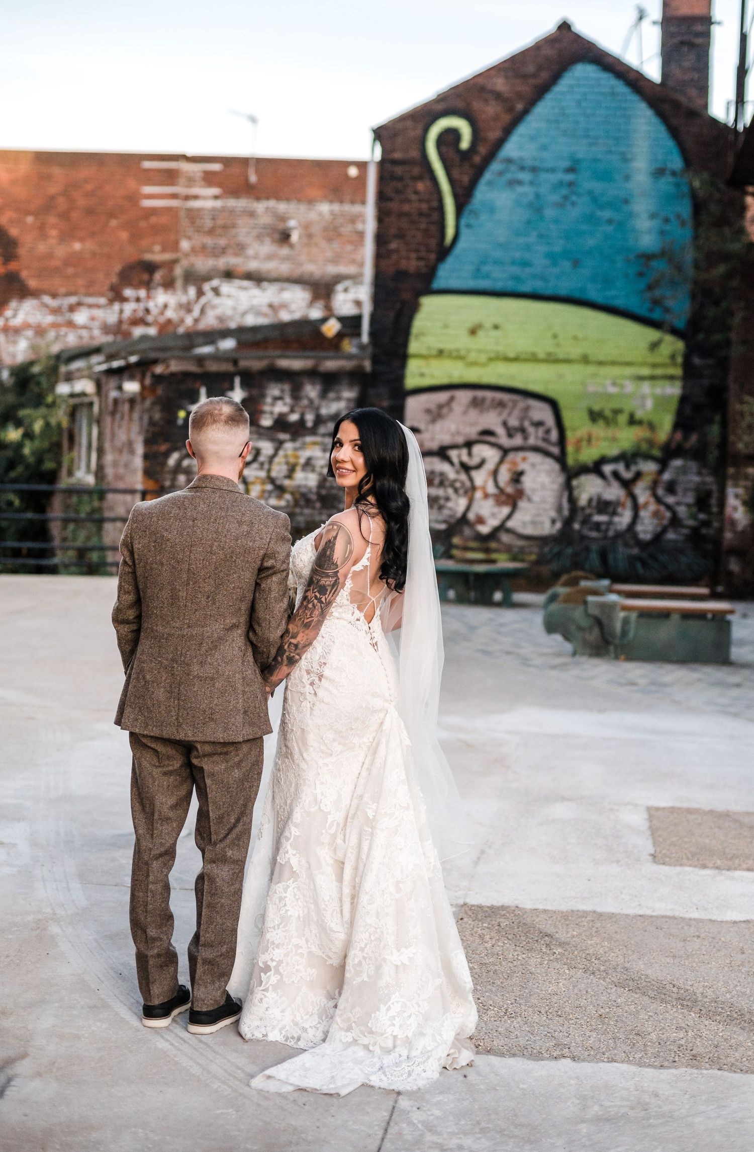 Bride in lace gown and groom in tweed suit stand before colorful street art mural.