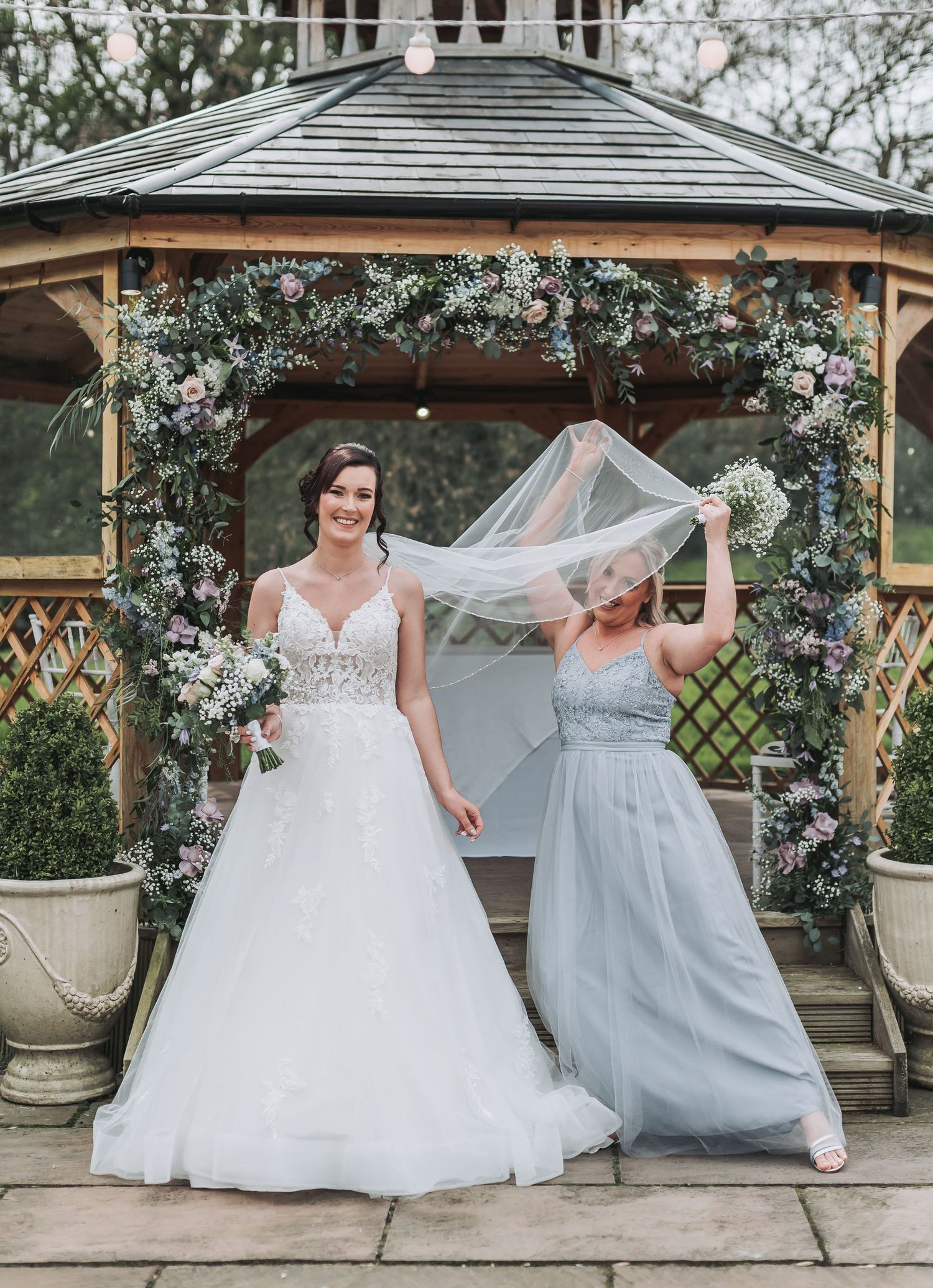 Two people in formal attire pose under a floral-decorated gazebo with flowing veil.