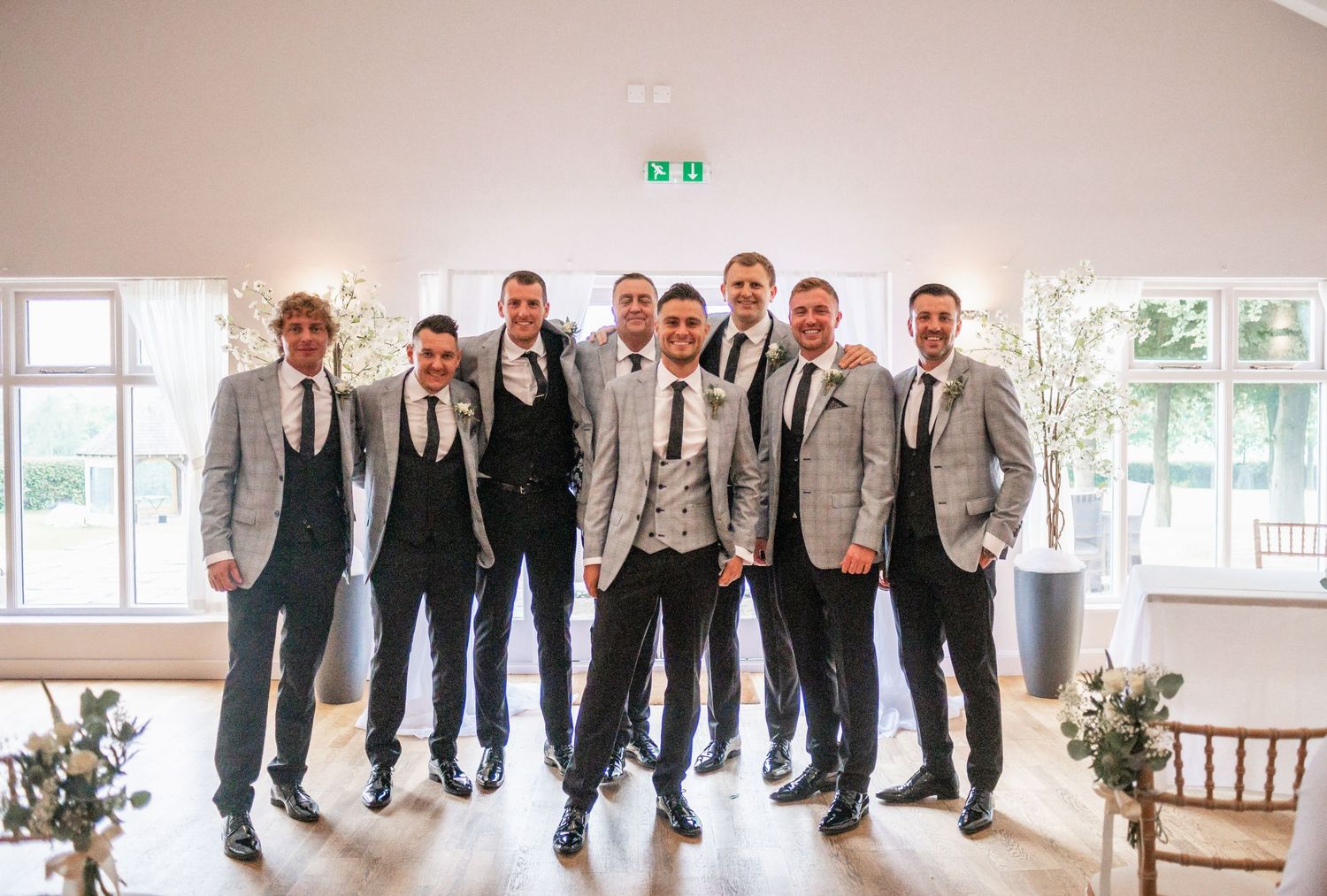Groomsmen in coordinated grey suits and black ties posing together in a bright venue.
