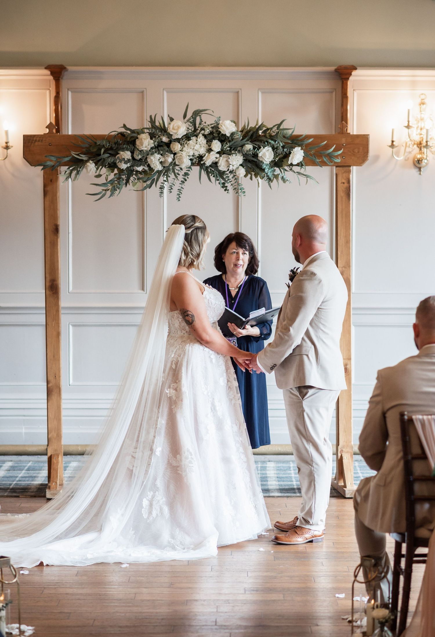 Wedding ceremony under wooden arch decorated with white floral arrangements.