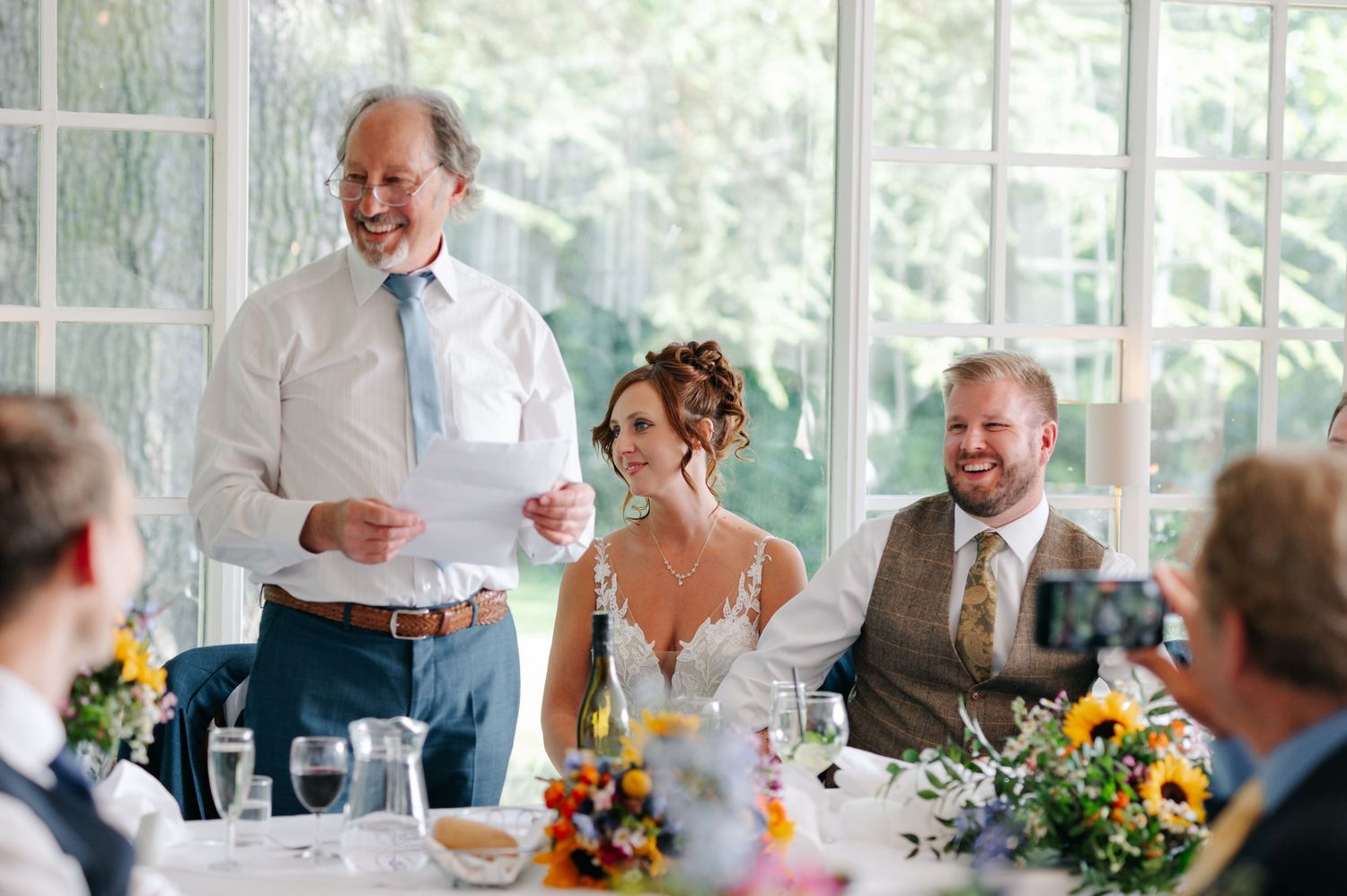 Wedding reception dinner scene with guests seated at decorated tables in a bright sunlit venue.