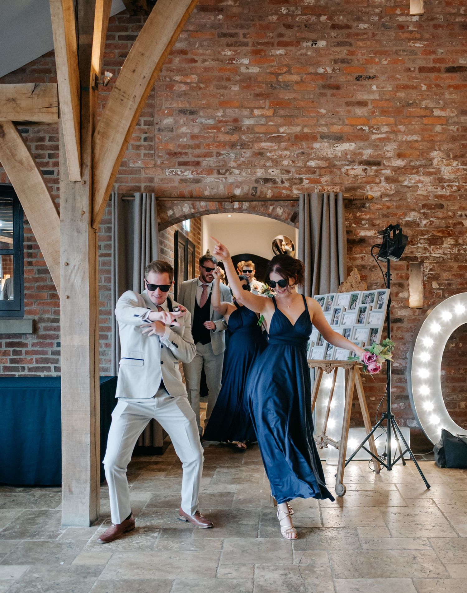 Wedding party strikes fun poses against rustic brick wall with illuminated letters.
