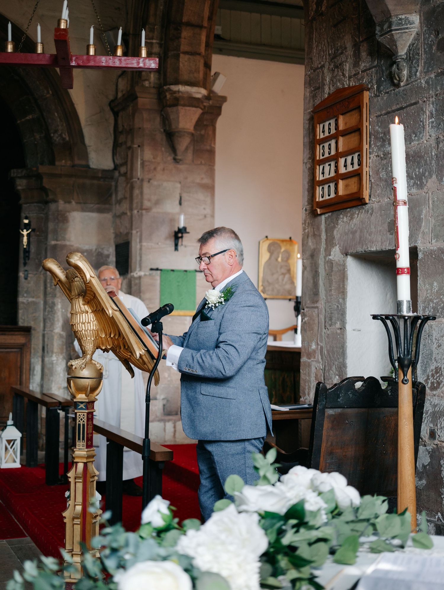 A person in a gray suit reads from a podium in a church decorated with white flowers.