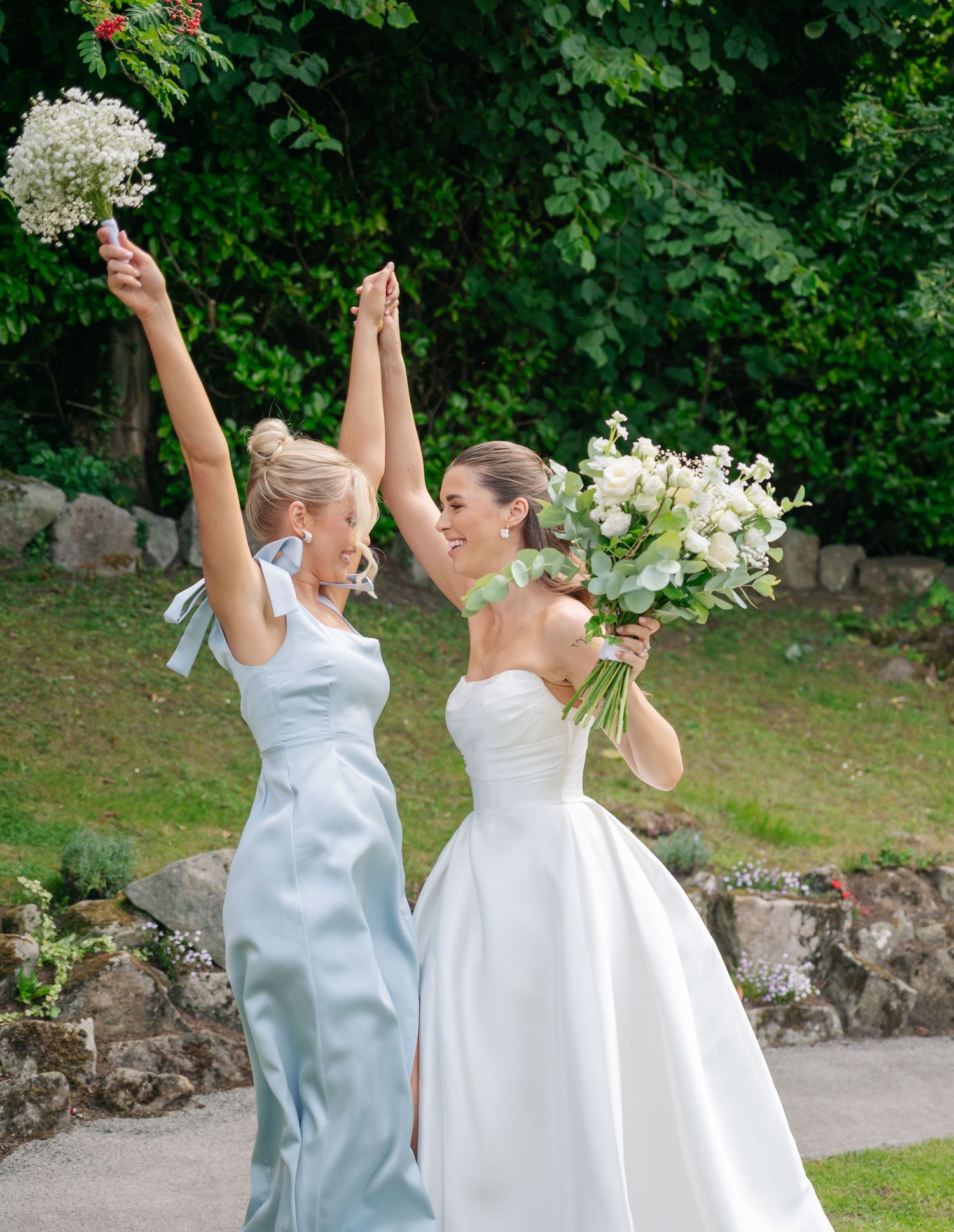 Two people in wedding attire celebrating joyfully outdoors with raised arms and bouquets against green foliage.