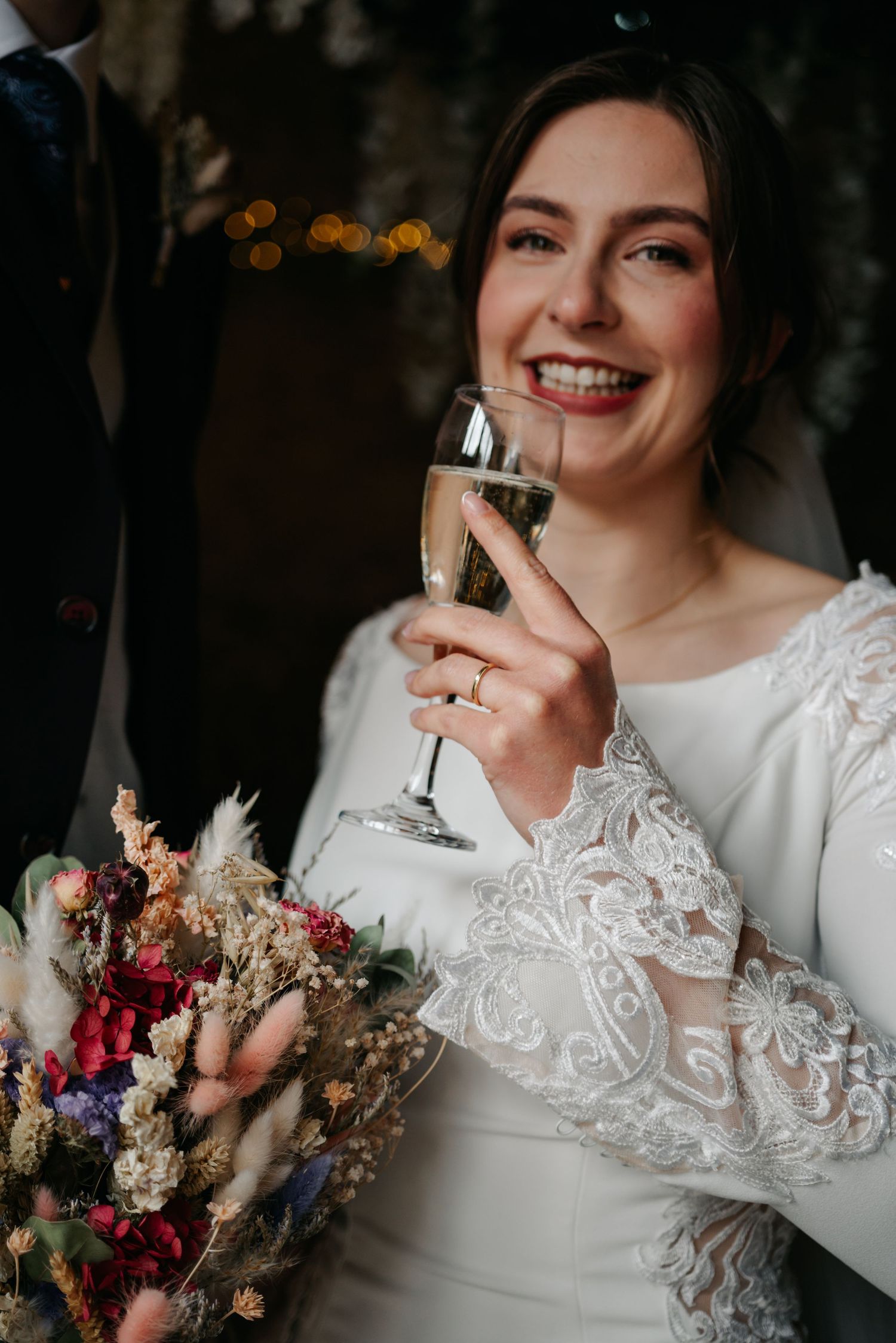 Close-up of bride in lace sleeve wedding dress holding champagne glass and colorful dried flower bouquet.