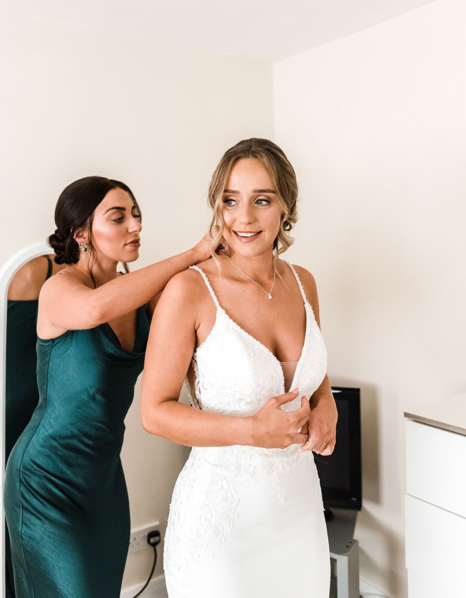 A bridesmaid in emerald green helps adjust jewelry on a bride in a white gown.
