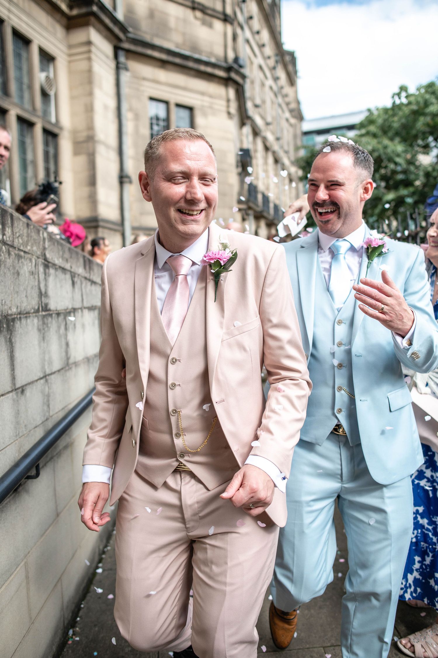 Wedding couple in coordinating suits walk together outside historic stone building.