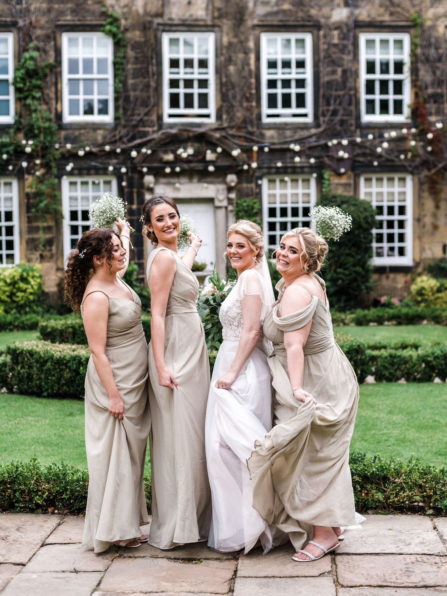 Bridesmaids in champagne dresses pose playfully on a stone patio of a historic mansion with string lights.