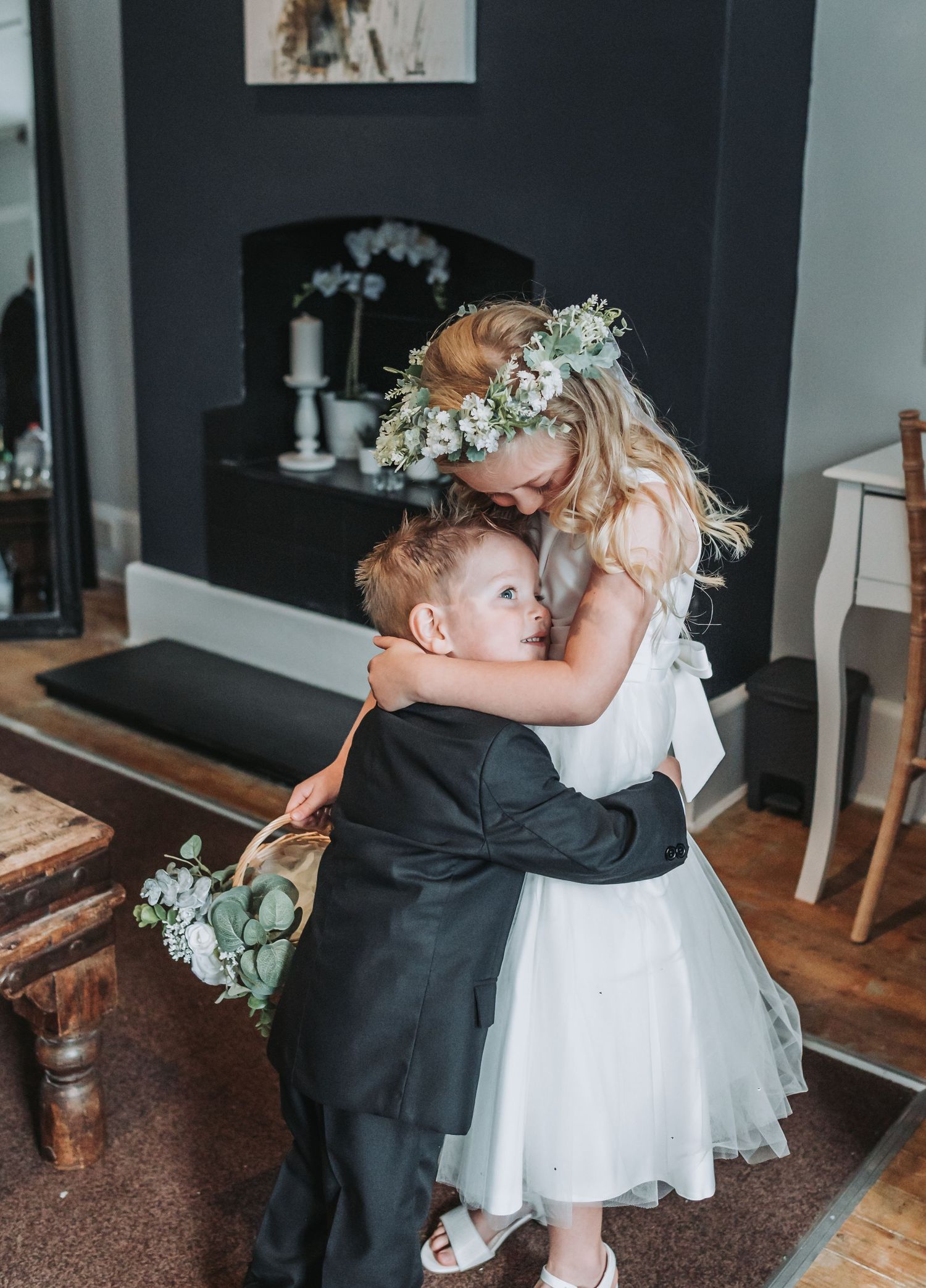 Sweet moment between flower girl in white dress and ring bearer in black suit sharing embrace.