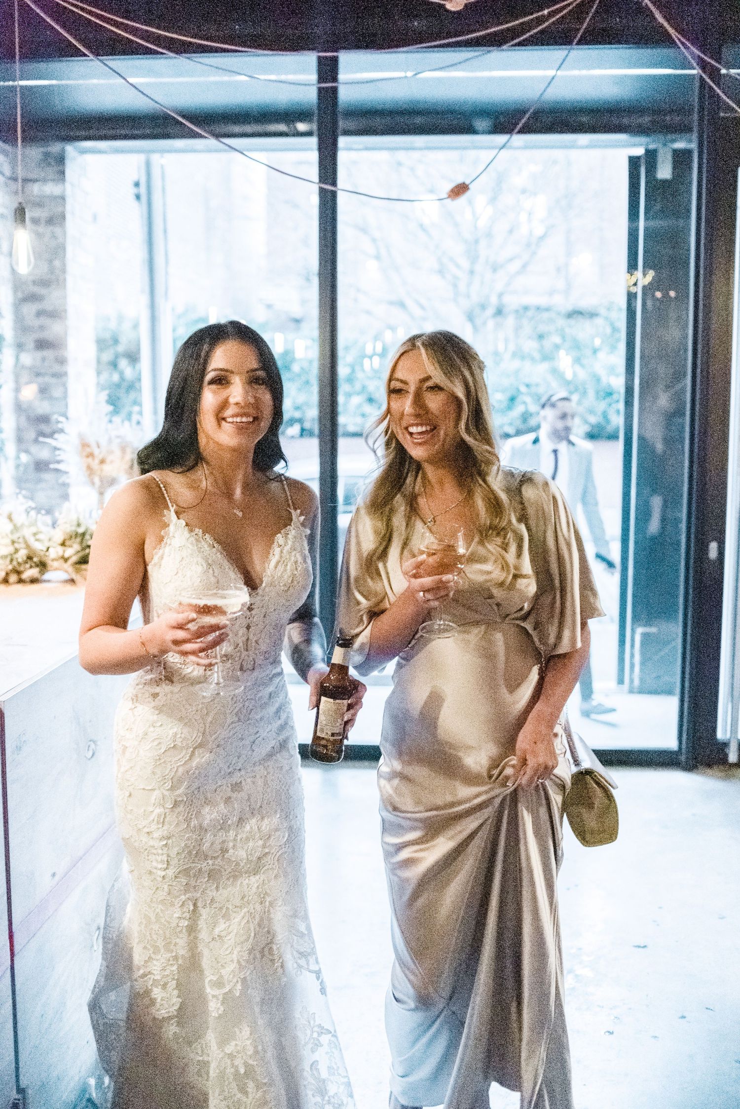 Two women in elegant dresses share drinks and laughter at wedding venue.