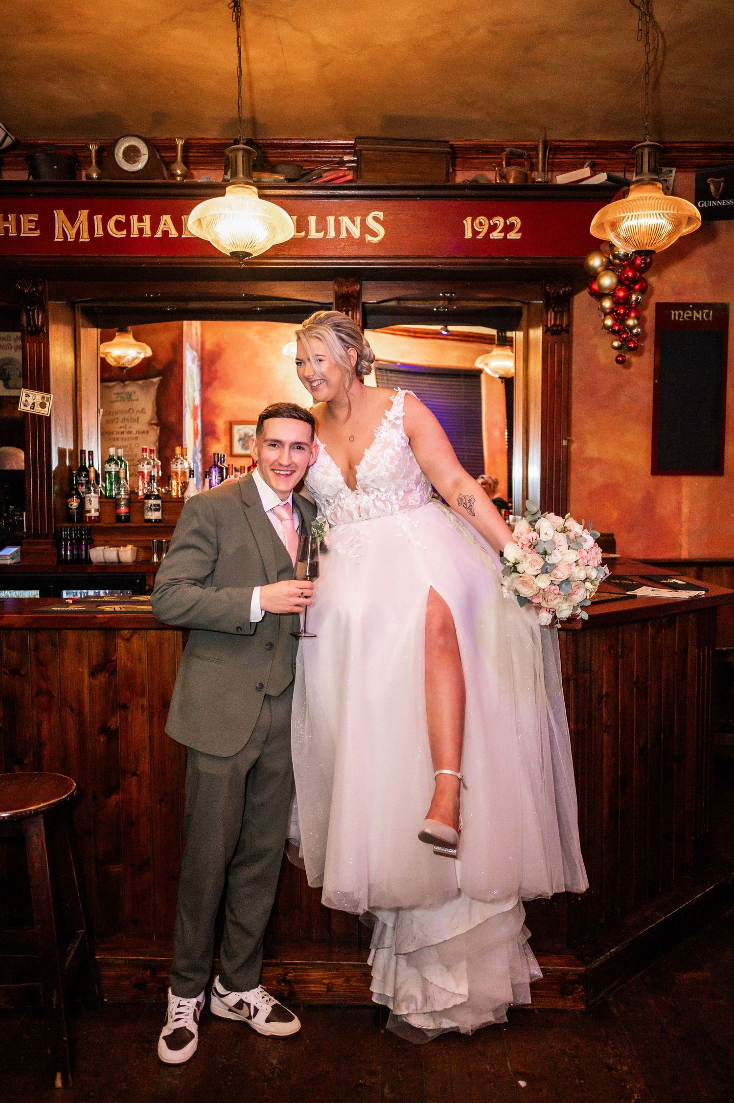 Newlyweds share a joyful moment at The Michael Collins pub with bride standing on bar in elegant white dress.