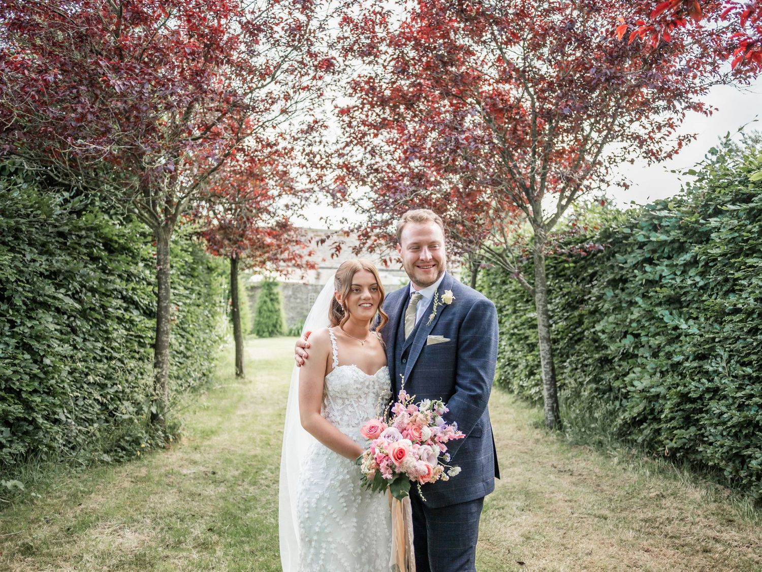 Wedding couple poses under red-leafed trees in manicured garden path.