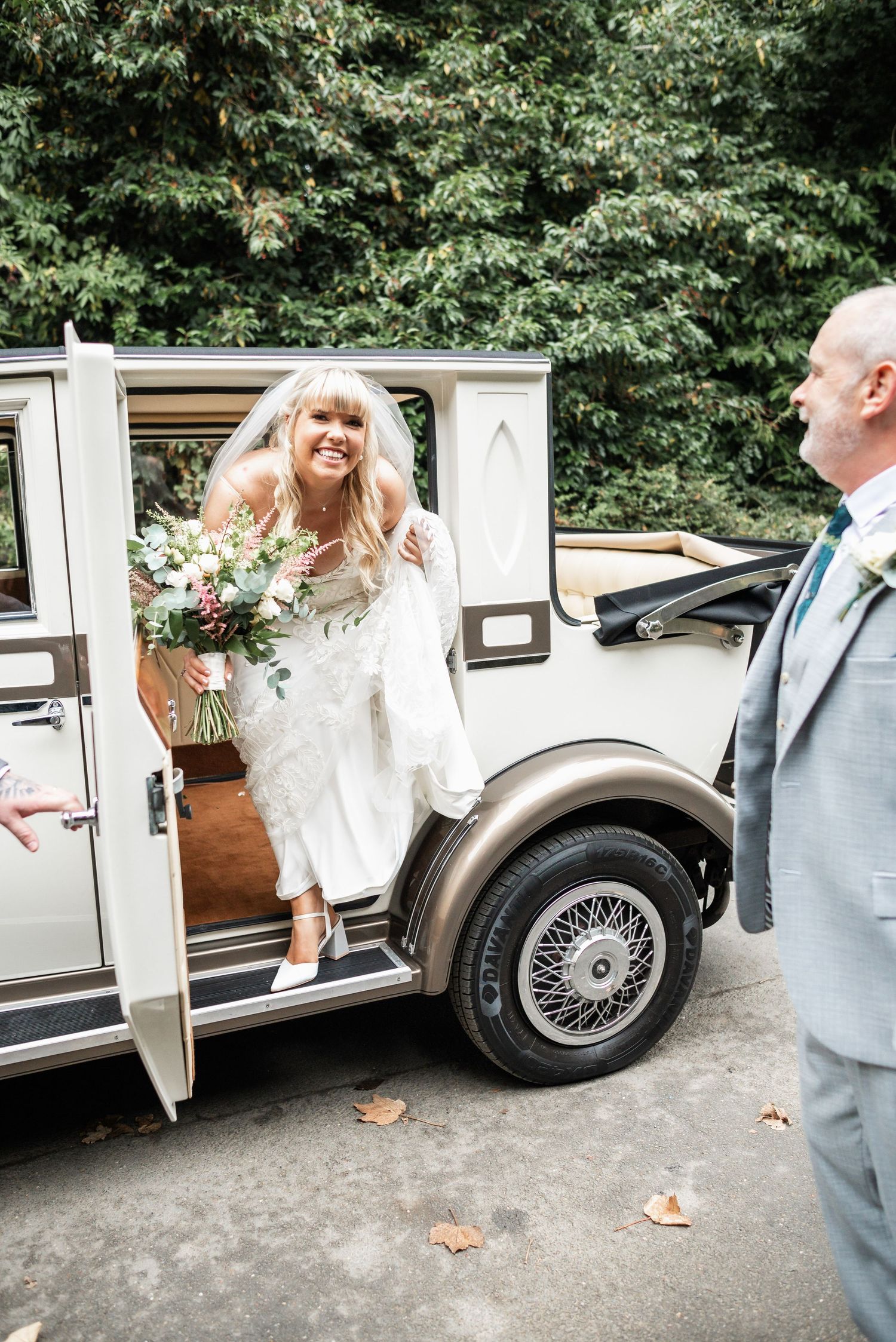 Bride in white dress emerges from classic vintage car while gentleman in grey suit greets her.