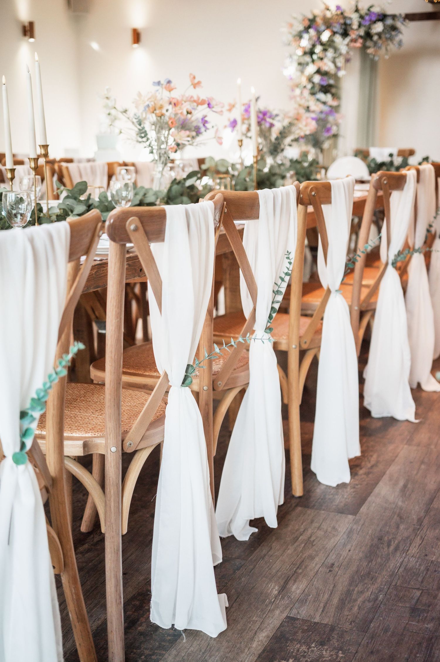 Wedding ceremony chairs decorated with flowing white fabric and floral accents in rustic venue.