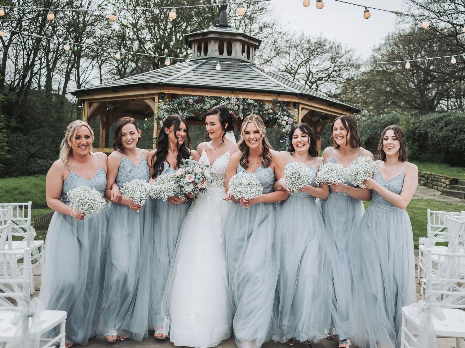 Bridal party in pale blue bridesmaid dresses poses together by a gazebo with string lights overhead.