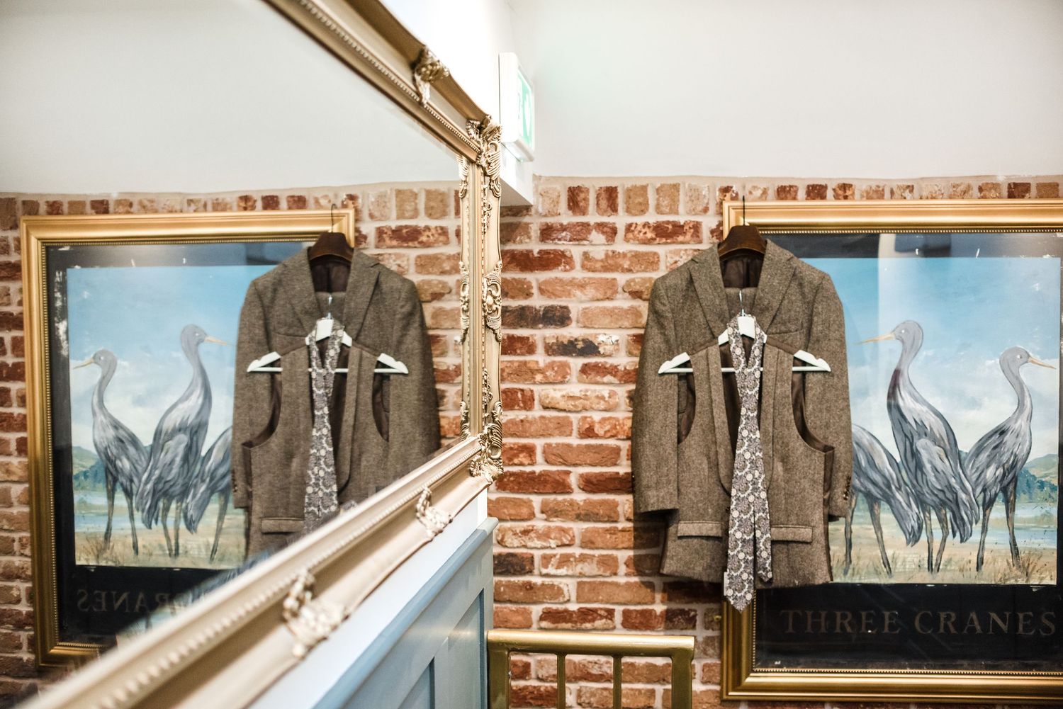 Two identical gray suits displayed on hangers against a brick wall with framed bird artwork.