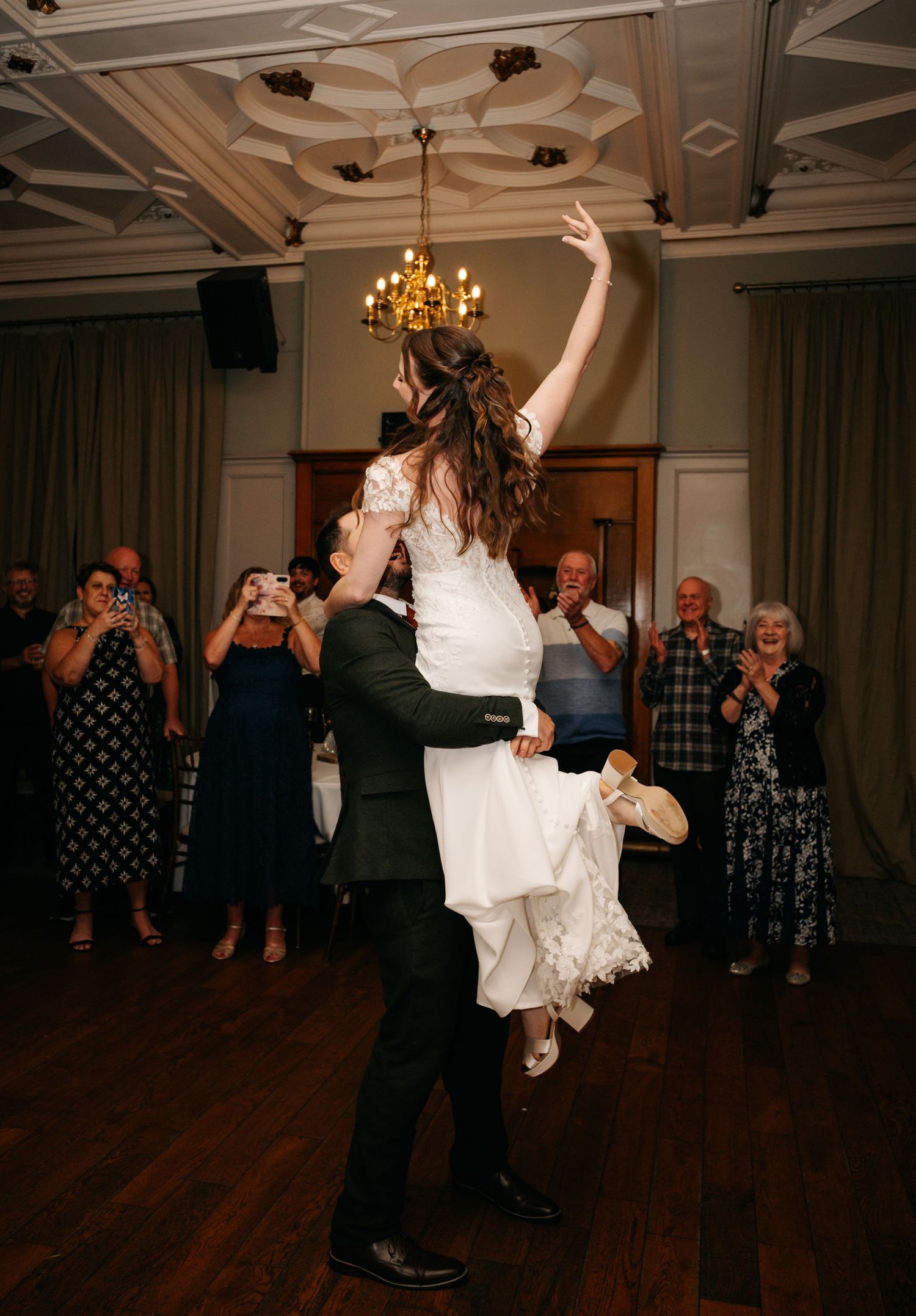 Groom lifts bride in a white lace gown high into the air during their first dance as smiling wedding guests watch and applaud.