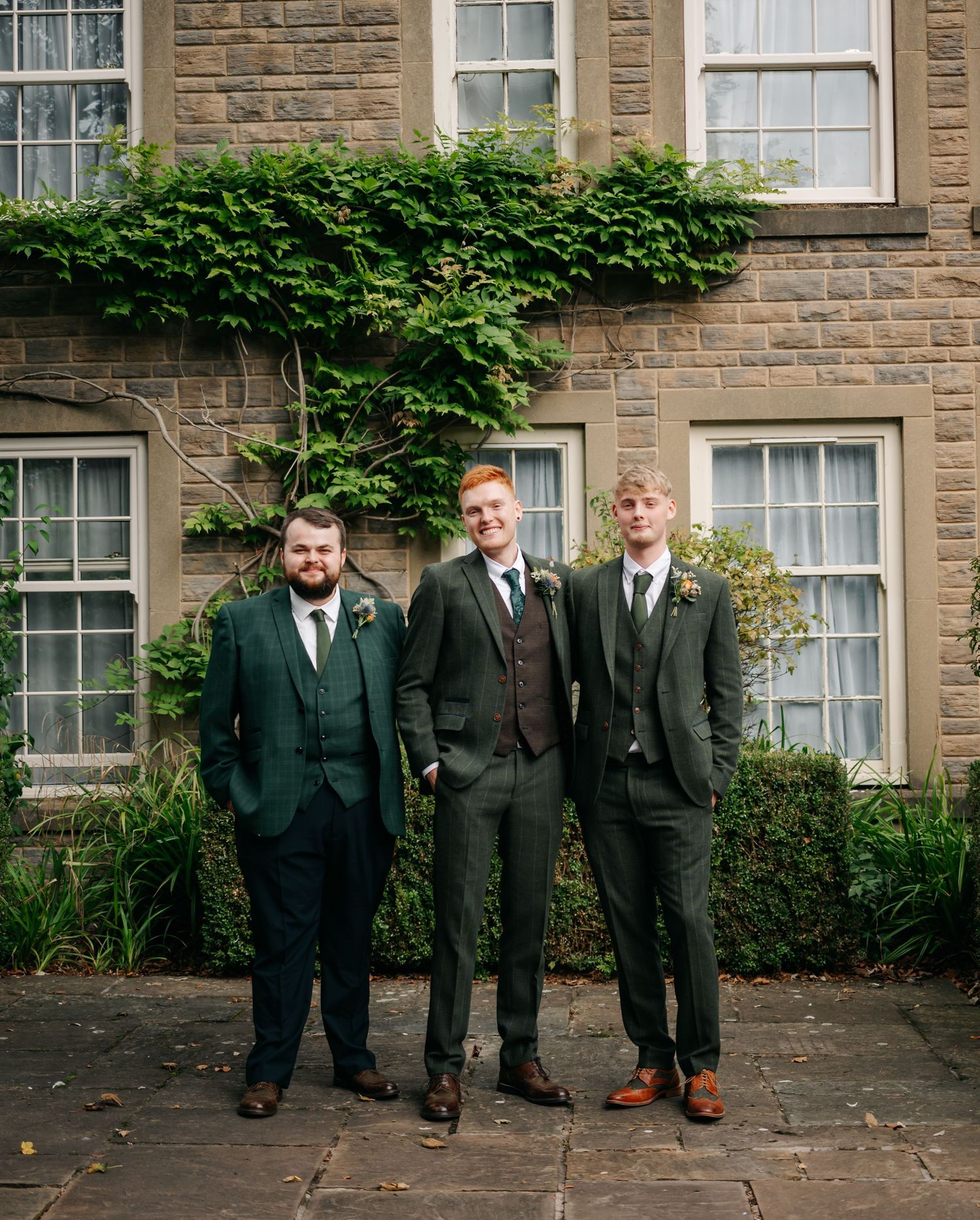 A group in dark suits poses outside a stone building with ivy-covered walls.