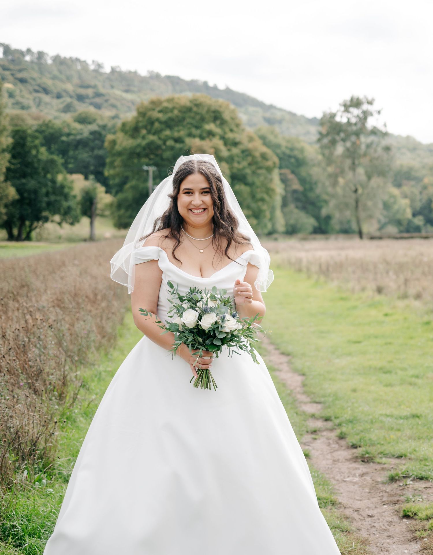 Bride in flowing white ballgown poses in scenic countryside meadow with rolling hills.