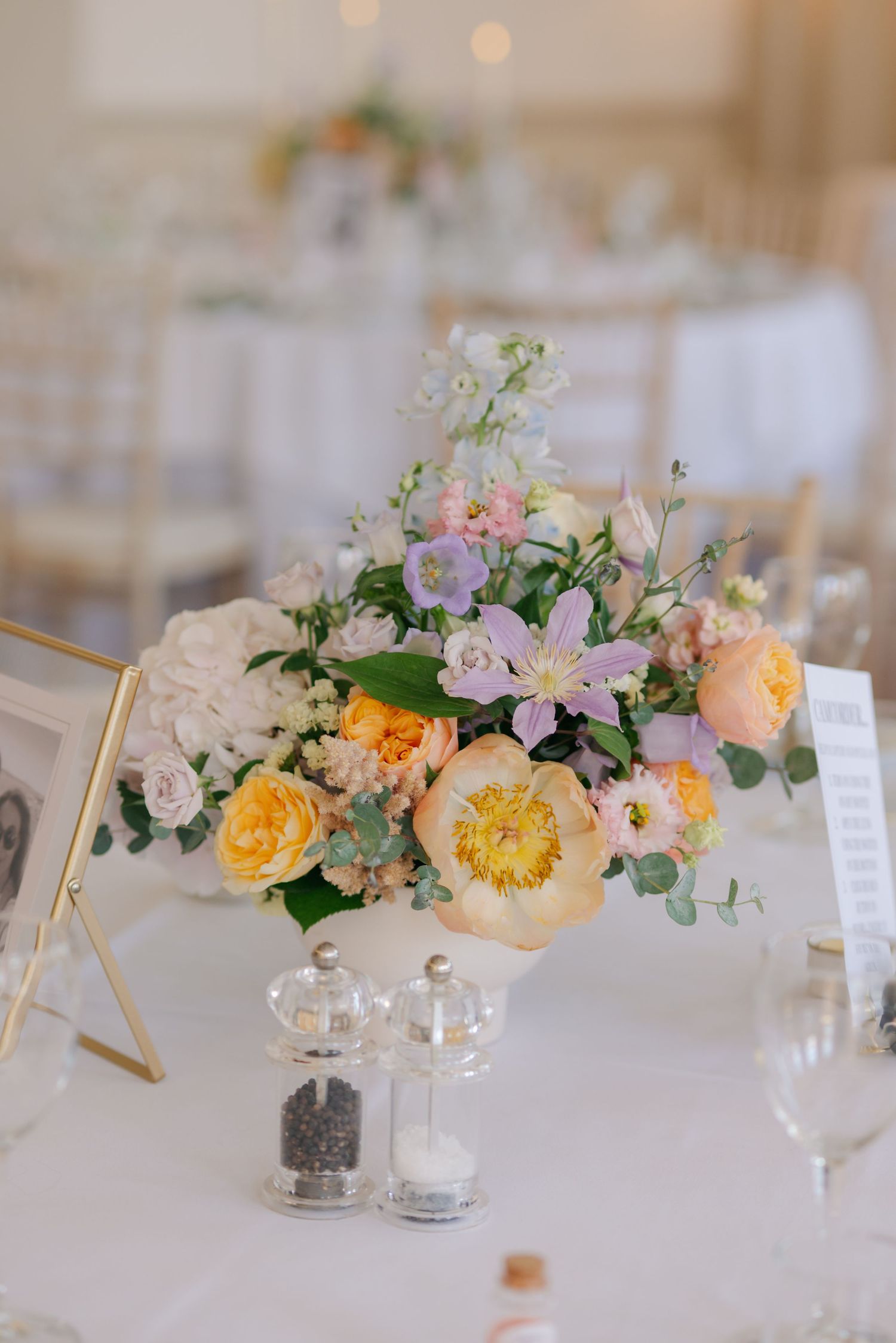 Elegant floral centerpieces with yellow and pink blooms decorate white tablecloths at a wedding reception.