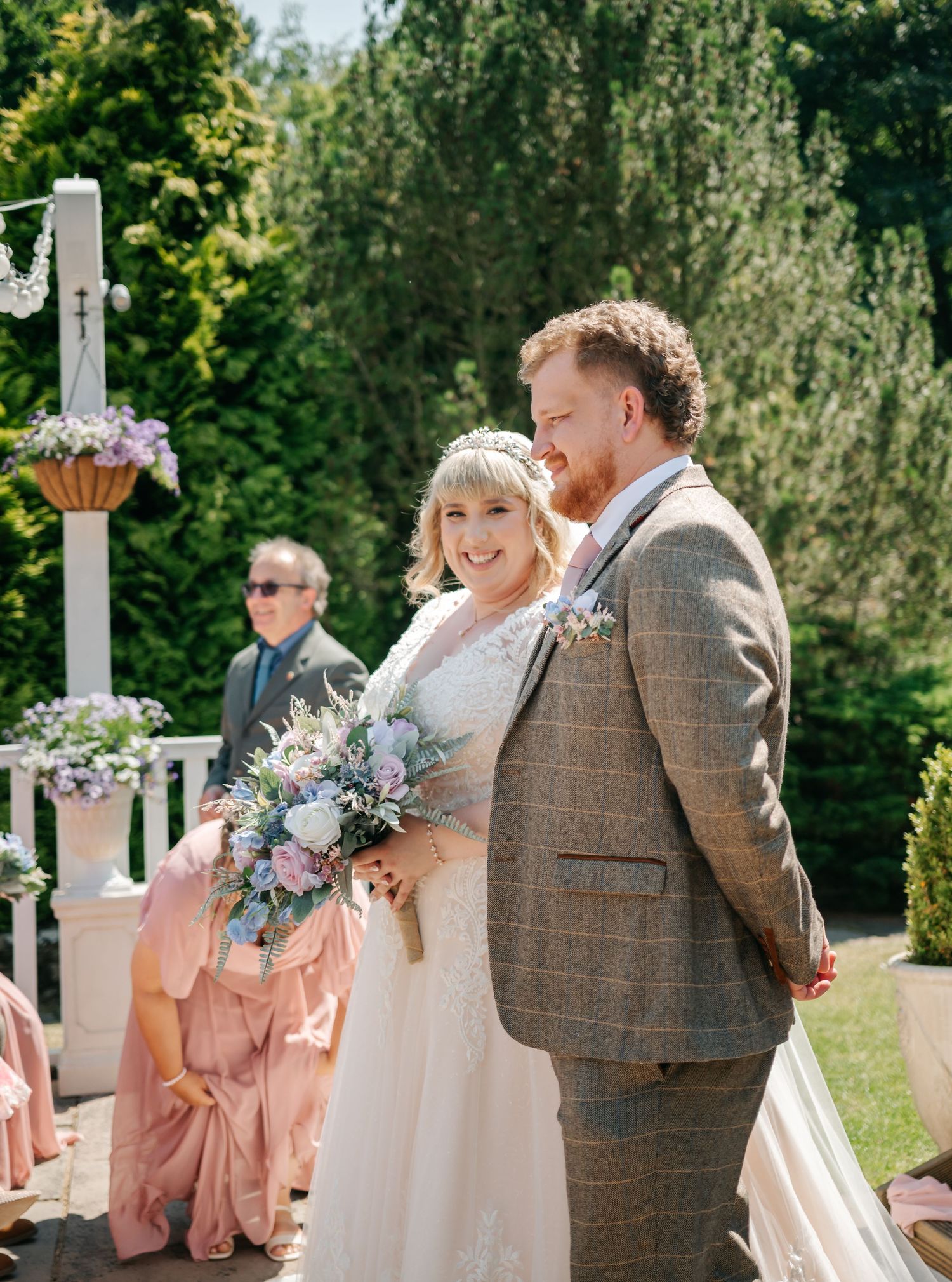 Bride and groom smiling during outdoor wedding ceremony, bride holding pastel bouquet, groom in brown tweed suit.
