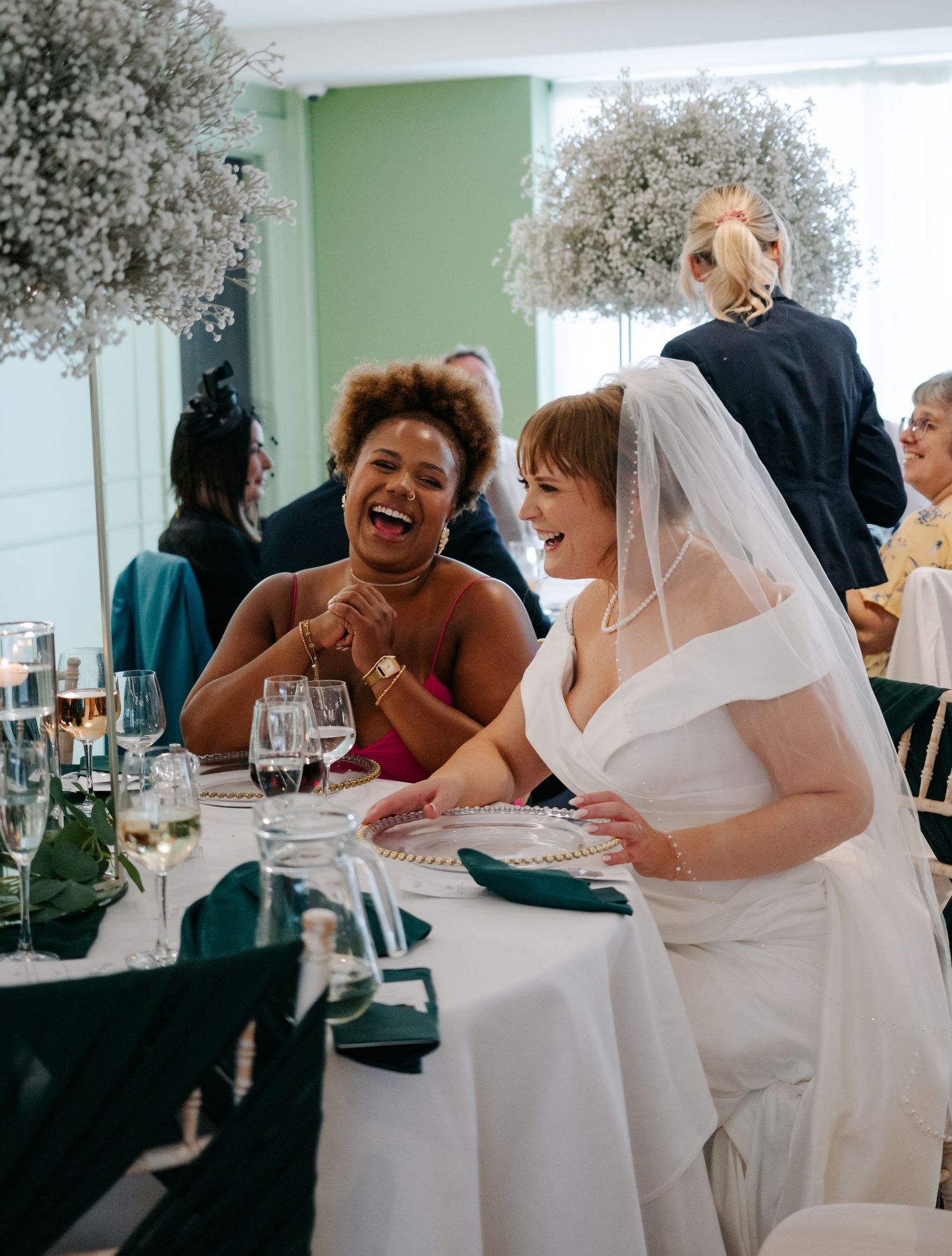 Wedding guests share joyful laughter around tables at an indoor reception with baby's breath decorations.