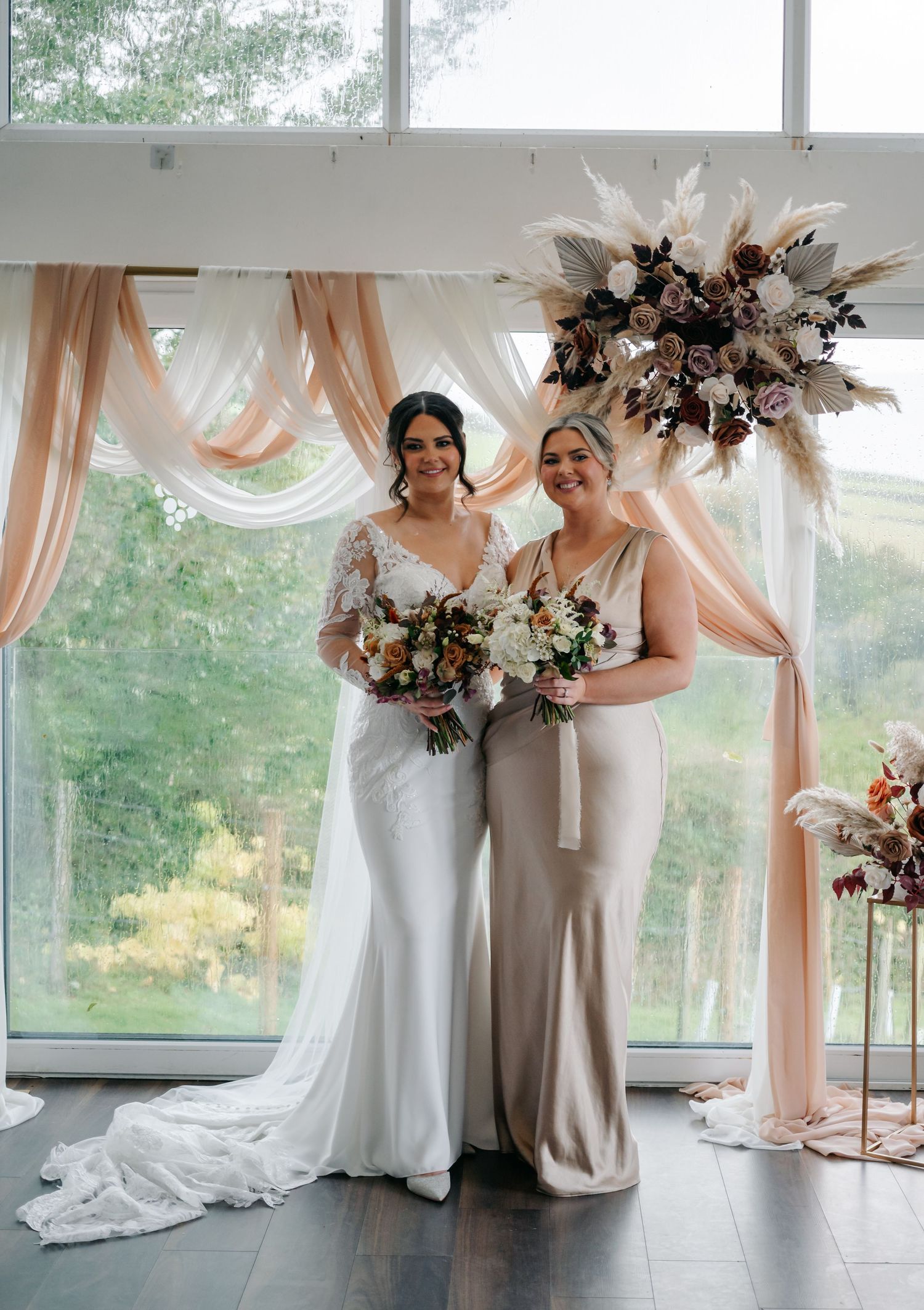 Wedding party members in neutral colors holding bouquets in a modern white venue with large windows.