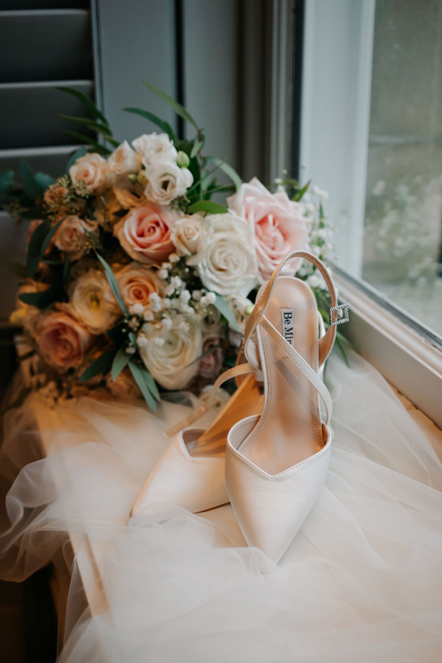 White bridal shoes and floral bouquet arranged on windowsill with natural light.