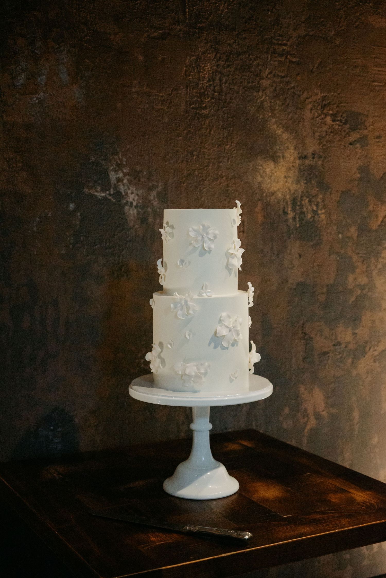 Elegant white wedding cake with delicate floral details displayed on a white pedestal against a dark rustic background.