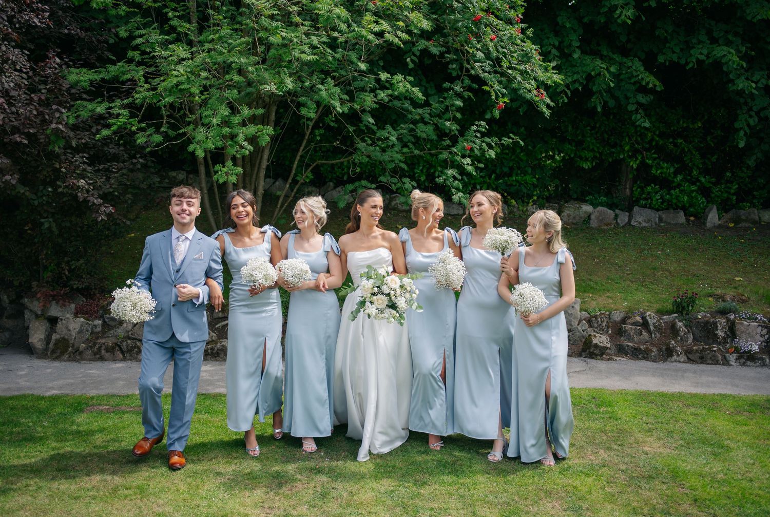 Bridesmaids in light blue dresses pose together on a grassy outdoor setting.