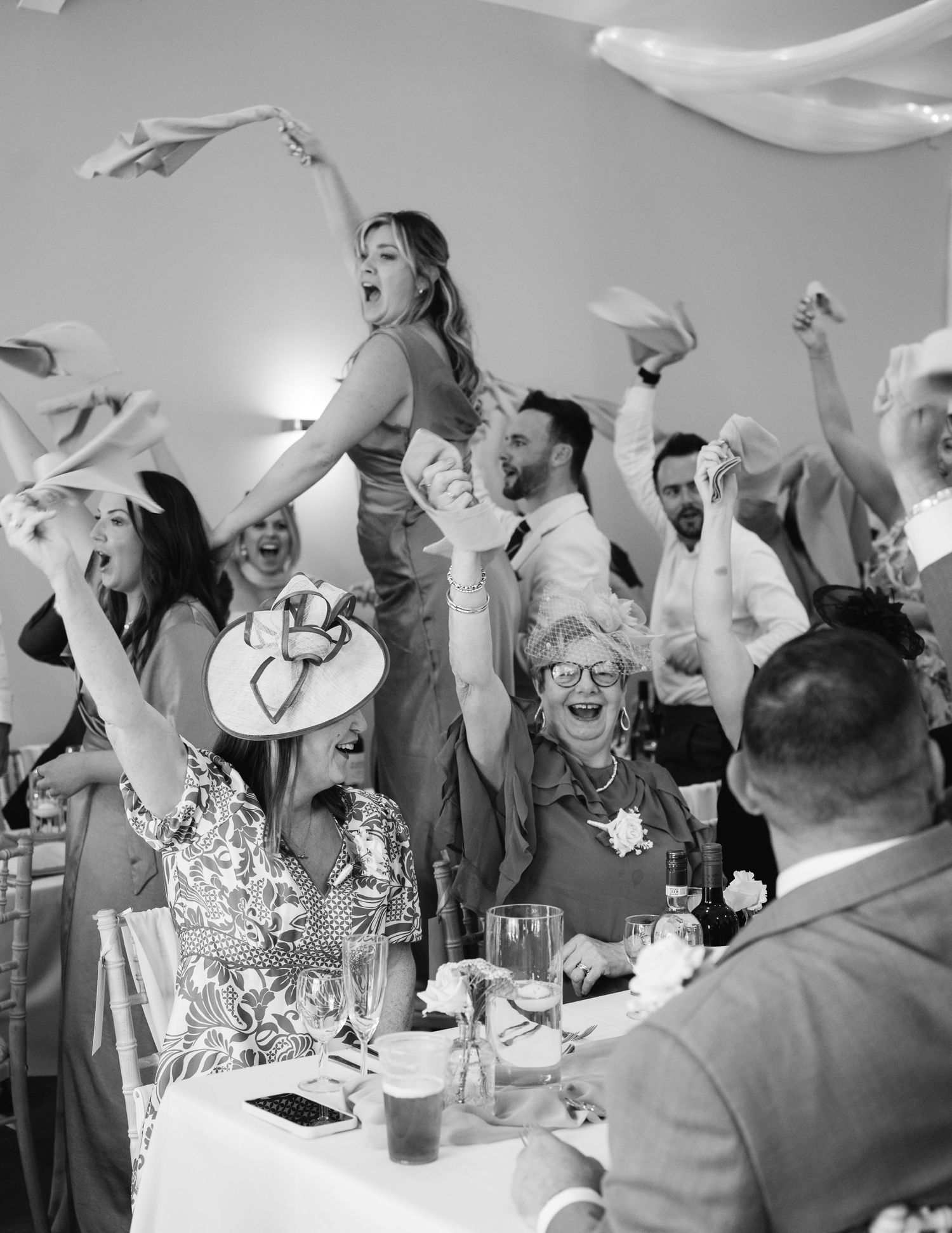 Black and white photo of joyful wedding guests celebrating, waving napkins and cheering at a reception dinner.