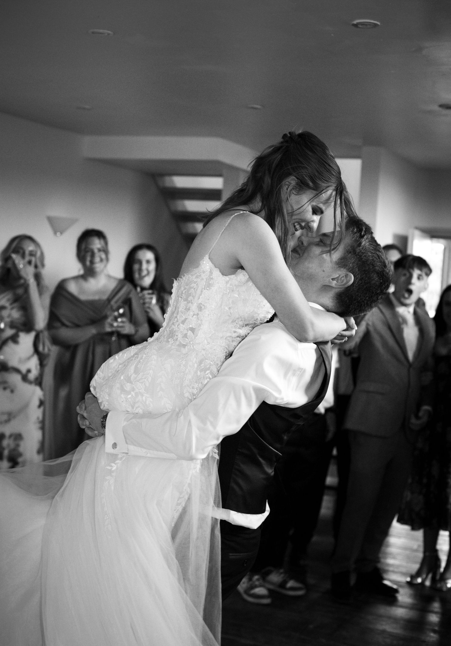 Groom lifts bride in wedding dress during first dance, guests react with joy in black and white photo.