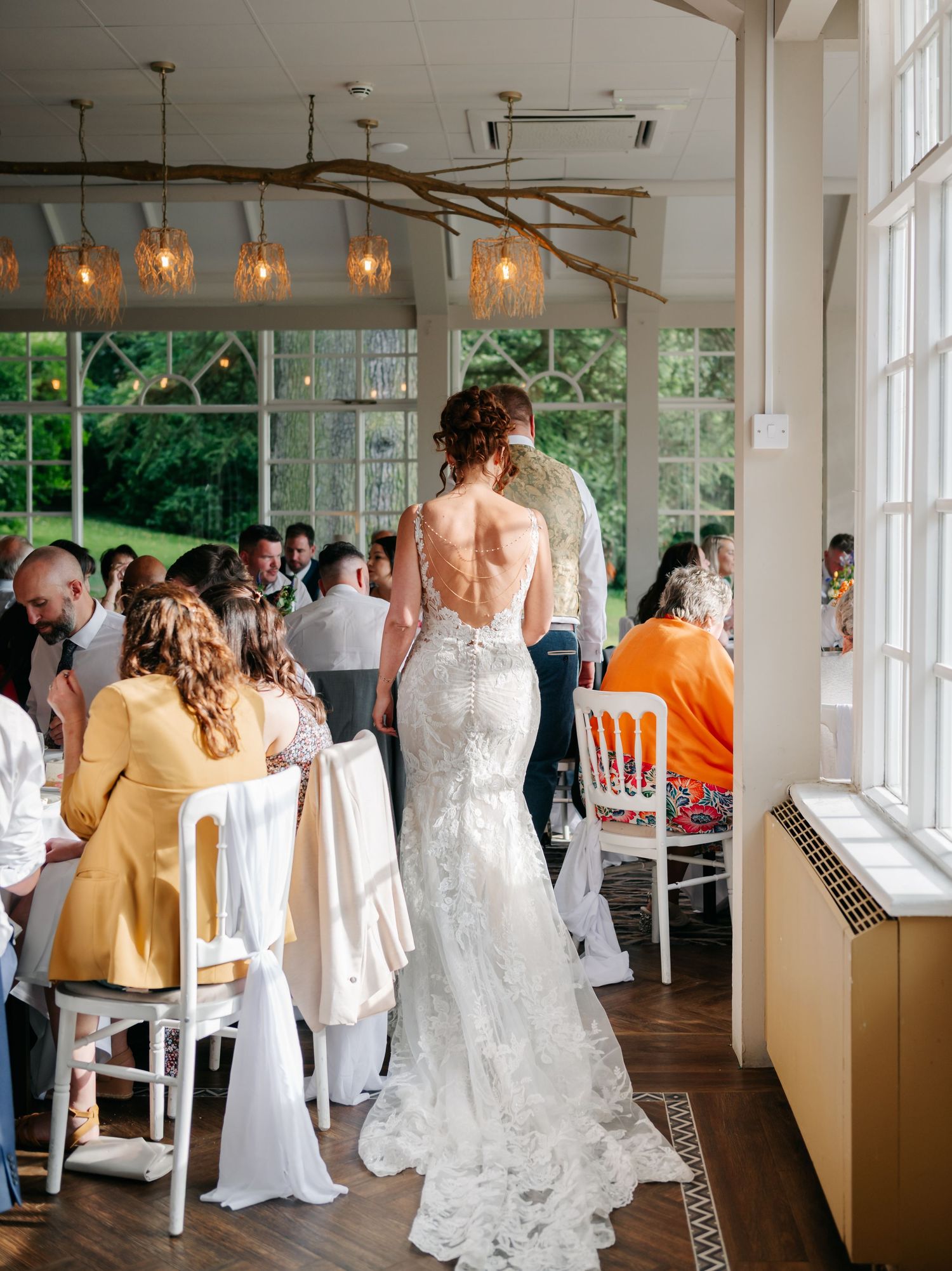A bride in a lace backless gown stands among seated guests in a bright reception venue with pendant lighting.